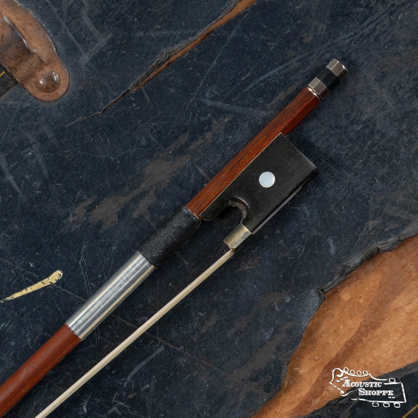 A close-up of the (Used) Horse Hair Fiddle Bow (5) by Used Items, featuring a wooden stick, silver winding, and black frog, resting on a dark textured surface with brown edges and the Acoustic Shoppe logo in the corner.