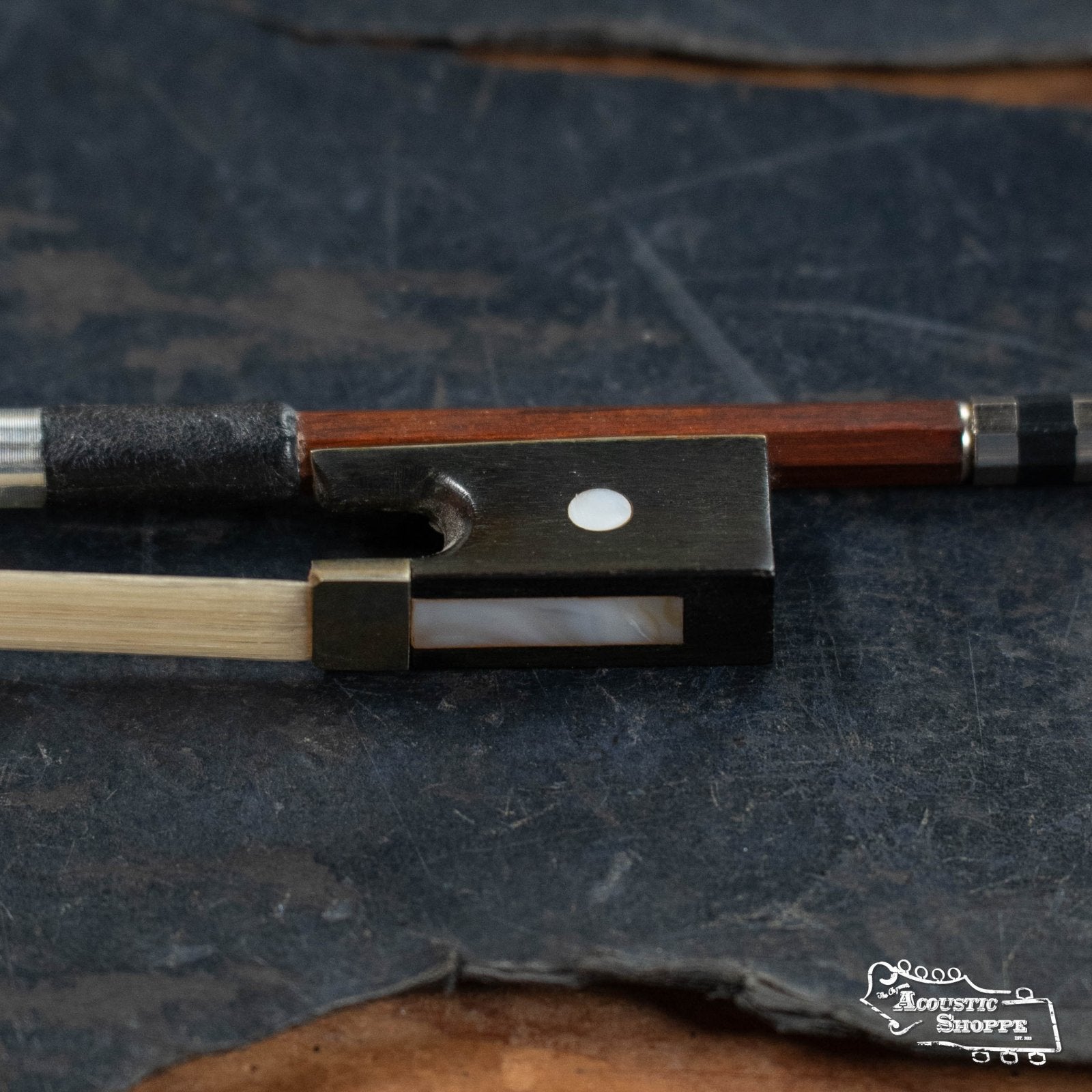 Close-up of the (Used) Horse Hair Fiddle Bow (5) by Used Items, featuring a frog with mother-of-pearl inlay, white dot, metal fittings, and a reddish-brown wood stick resting on a dark textured surface.