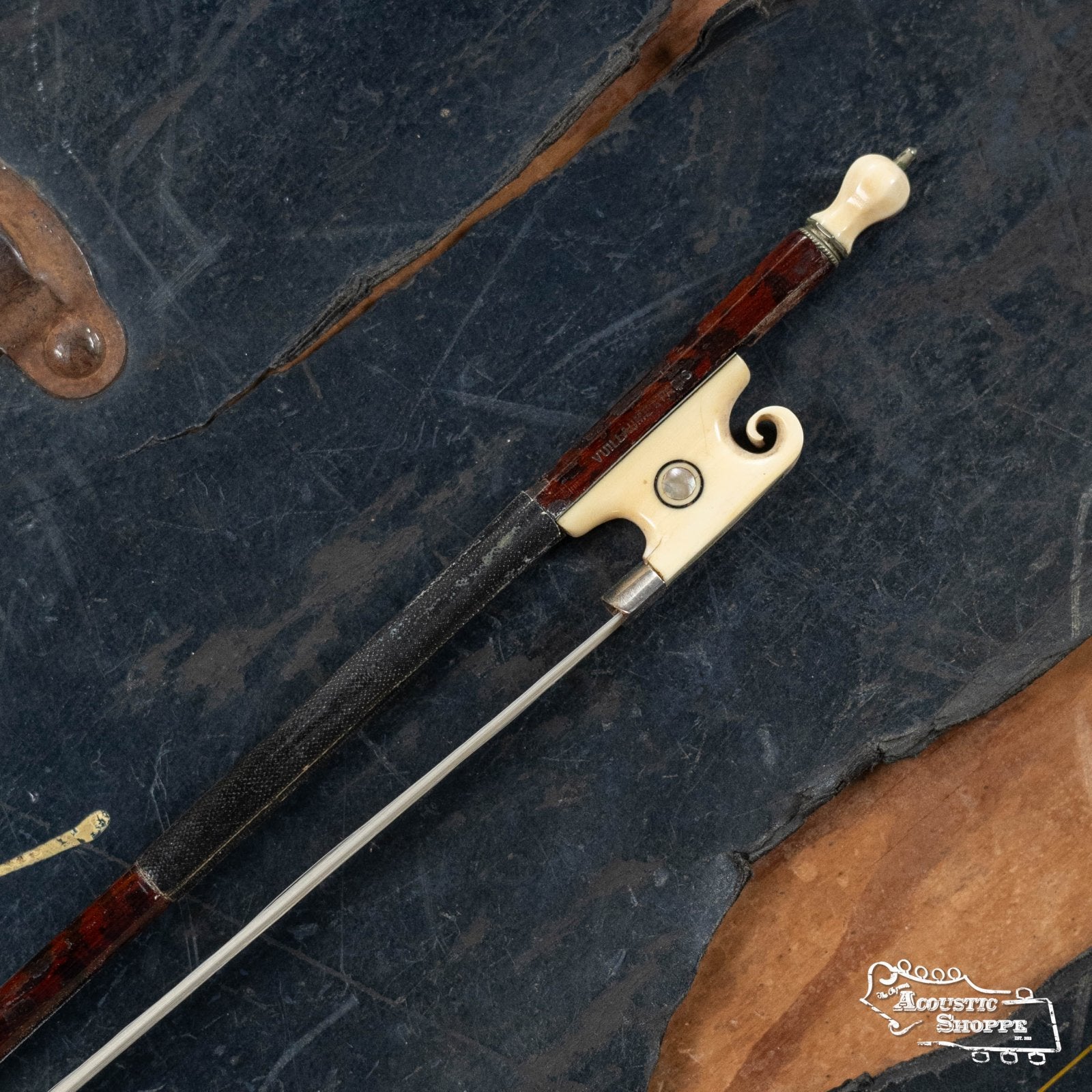 A close-up of the (Used) Horse Hair Fiddle Bow (2) by Used Items, featuring an ornate ivory frog and tip, lies on a textured dark surface with brown edges. The bow hair and winding stand out, with The Acoustic Shoppe logo in the corner.