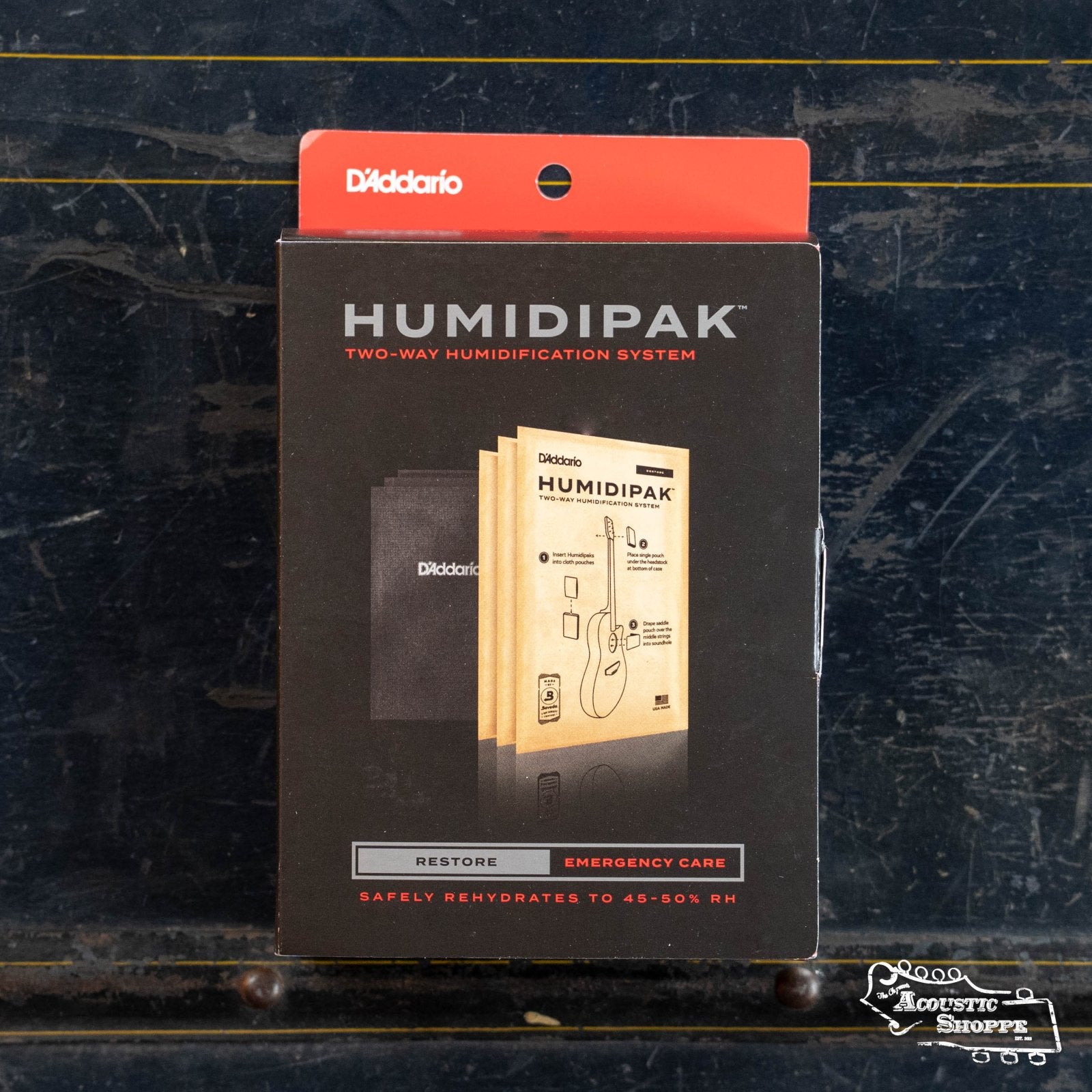 A D'Addario Humidipak Restore Kit PW-HPK-03 box with Two-Way Humidification System branding, shown upright against a dark, textured background, highlights advanced guitar humidity control and features a small shop logo in the corner.