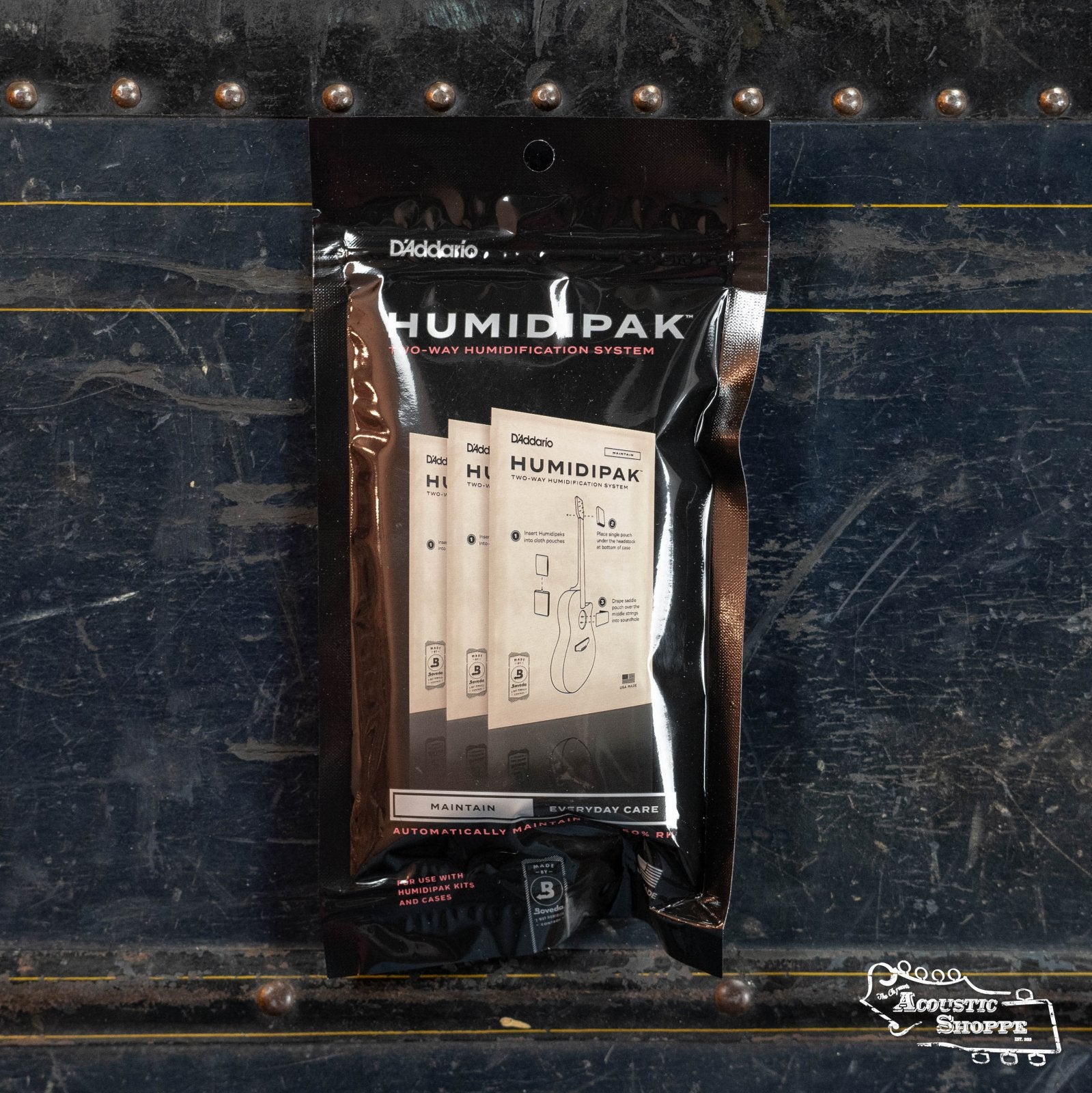 A pack of DAddario PW-HPRP-03 Humidipak 3-Packs Replacement Packets stands upright against a dark, textured case with metal trim. The Acoustic Shoppe logo is in the bottom right corner. Ideal for guitar humidity control.