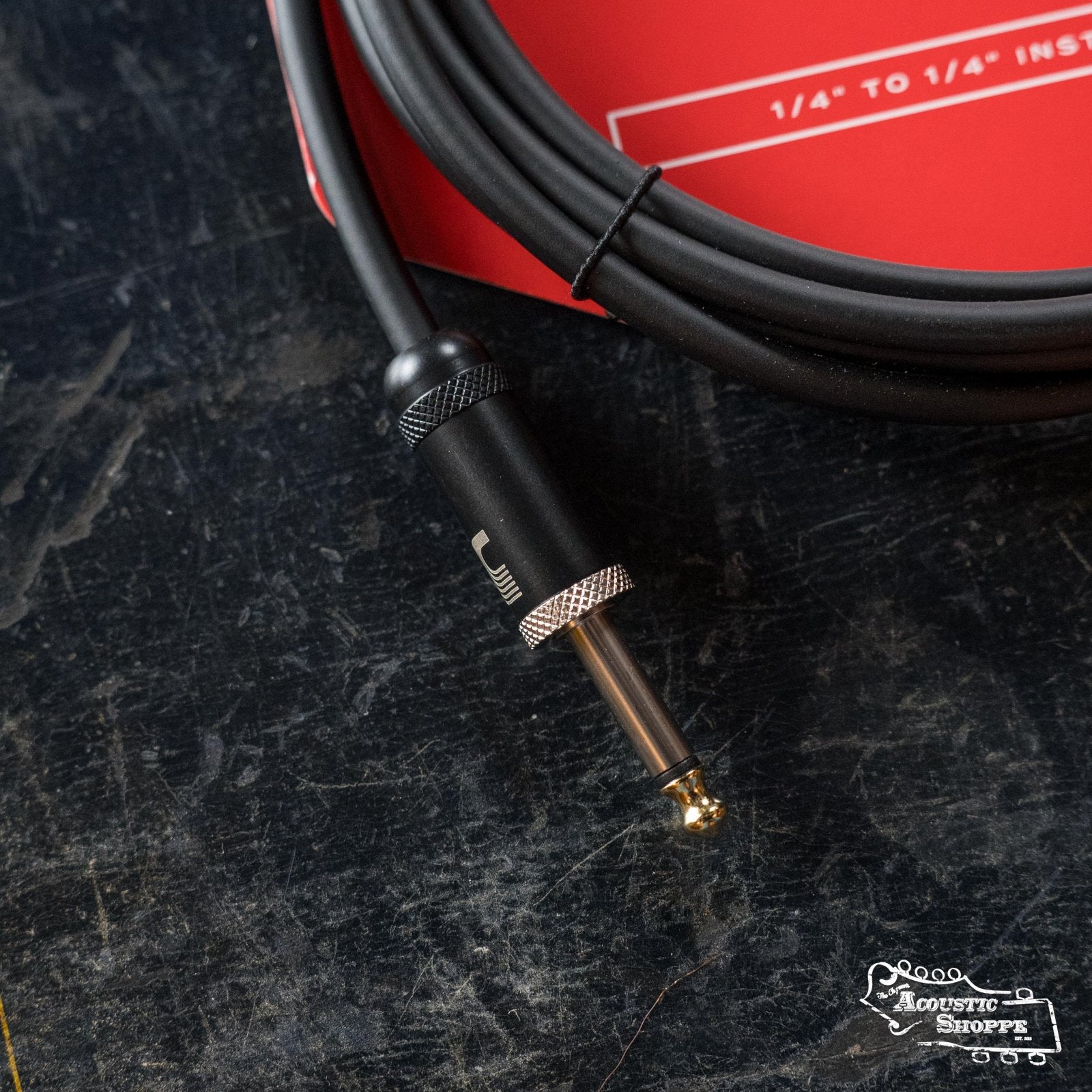 A close-up of a D'Addario American Stage Instrument Cable (10ft, PW-AMSG-10) lies on a dark surface, with part of a red label in the background and a small Acoustic Shoppe logo at the bottom right.