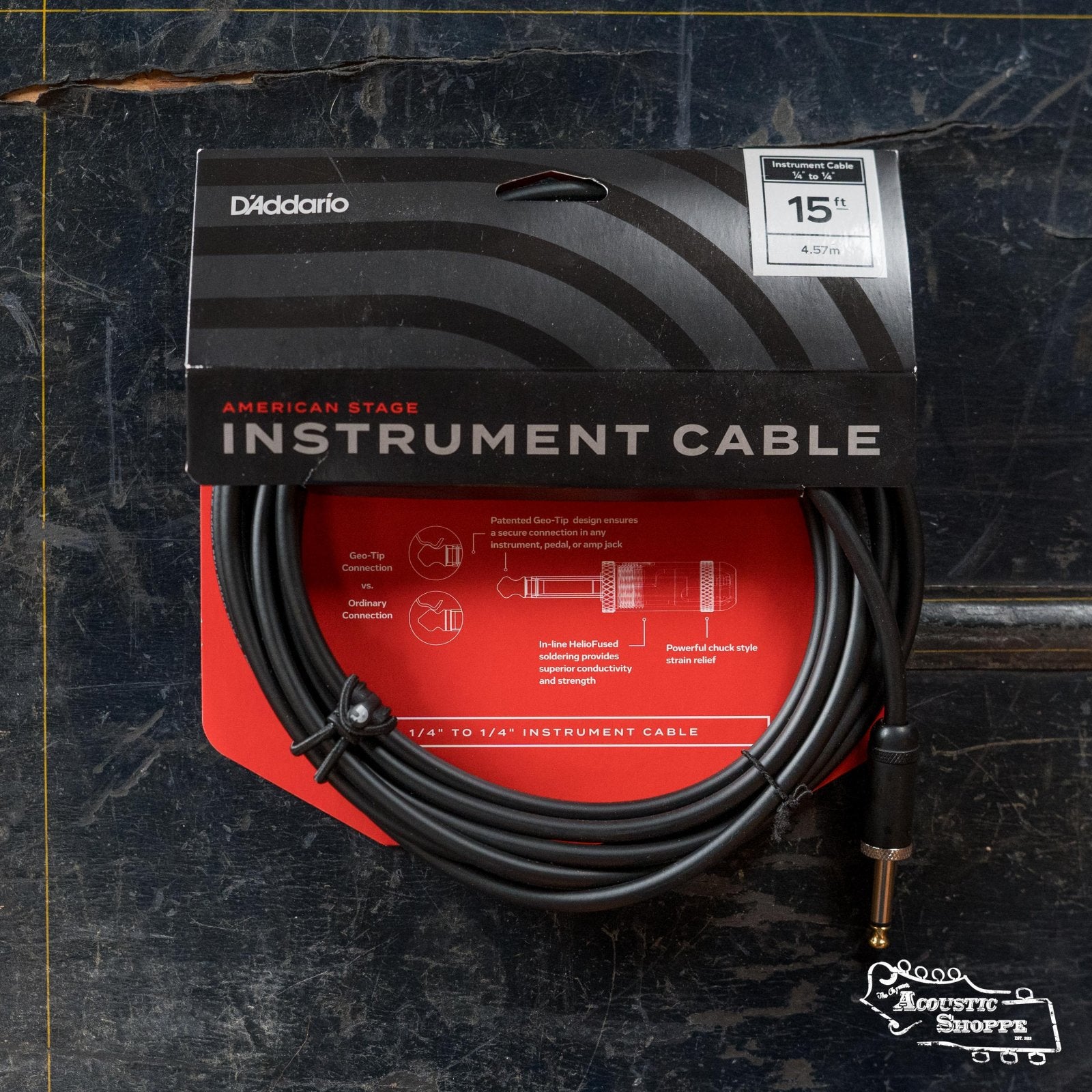 A D'Addario PW-AMSG American Stage Instrument Cable (1/4" to 1/4", 15 ft) by D'Addario, coiled on red and black packaging, rests on a worn dark surface with the Acoustic Shoppe logo in the bottom right.