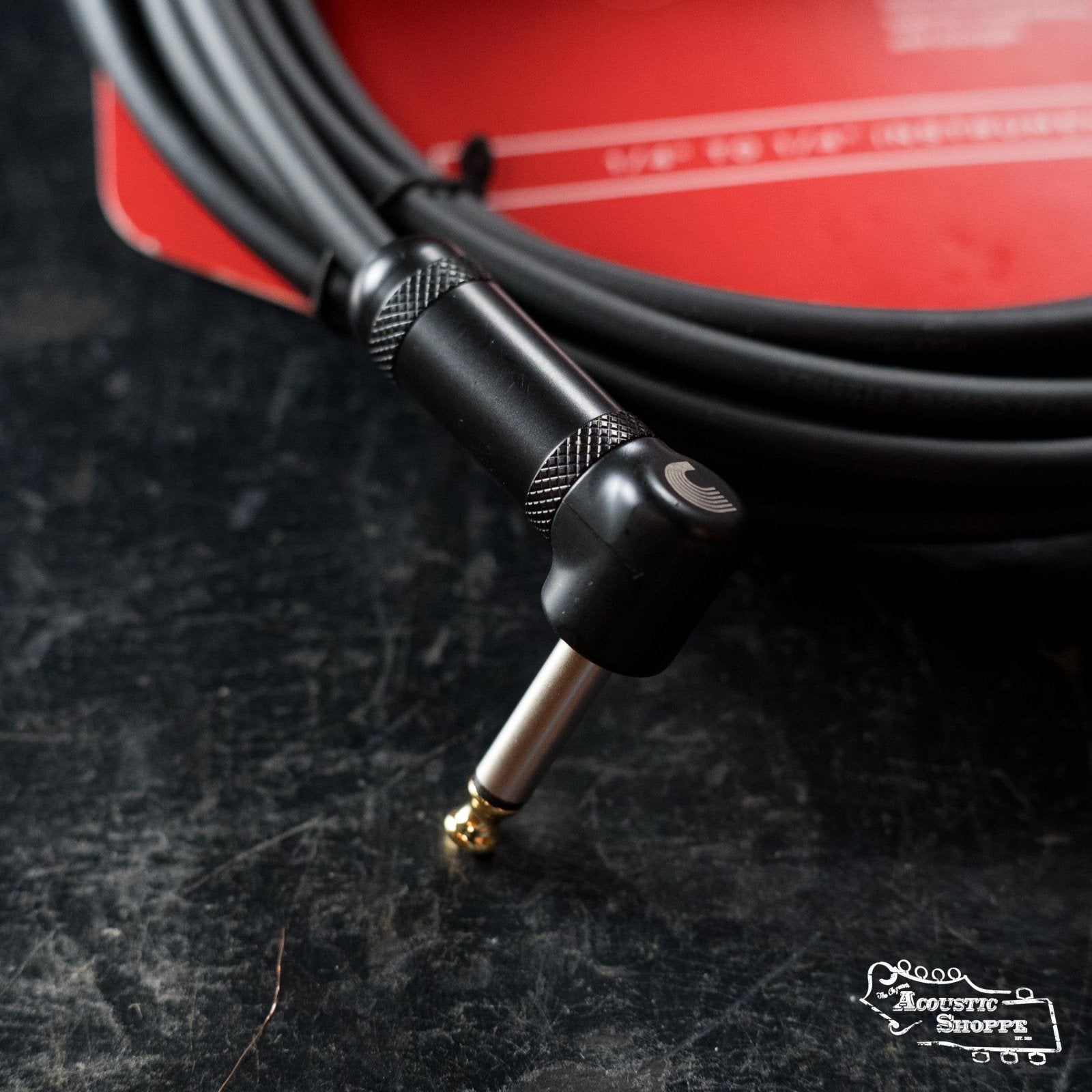 A D'Addario American Stage Instrument Cable Right to Straight 20ft (PW-AMSGRA-20) lies coiled on a dark surface; a red label is partly visible behind, with the Acoustic Shoppe logo in the lower right corner.