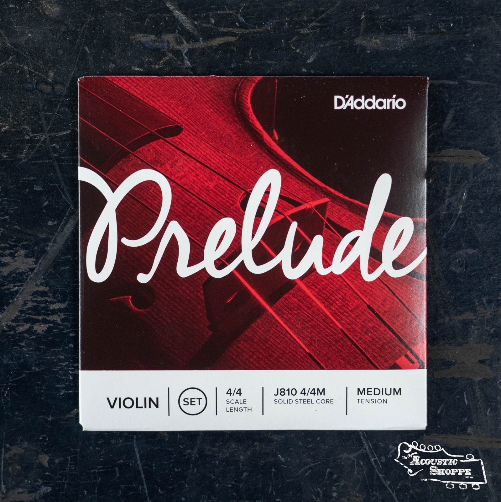 A DAddario Prelude Violin String Set 4/4 (J810) package in red and white, with a violin image and labels for solid steel core and medium tension, is placed on a dark surface.