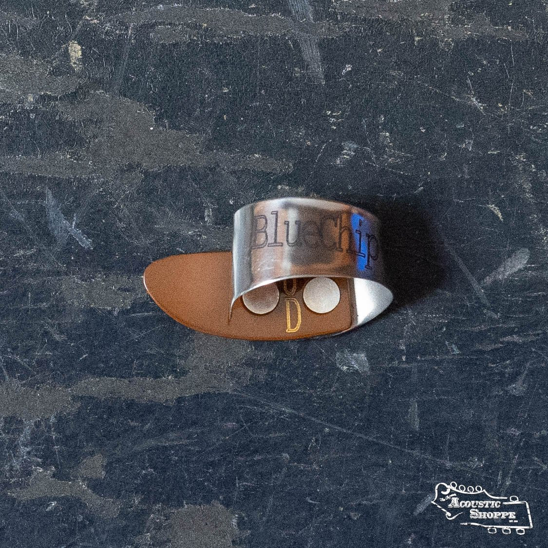 A BlueChip Picks (BCT-JDS) thumb pick by BlueChip, featuring a metal band and brown tip, sits on a textured dark surface. "BlueChip" is printed on the band above two white dots, making it perfect for precise acoustic guitar playing.