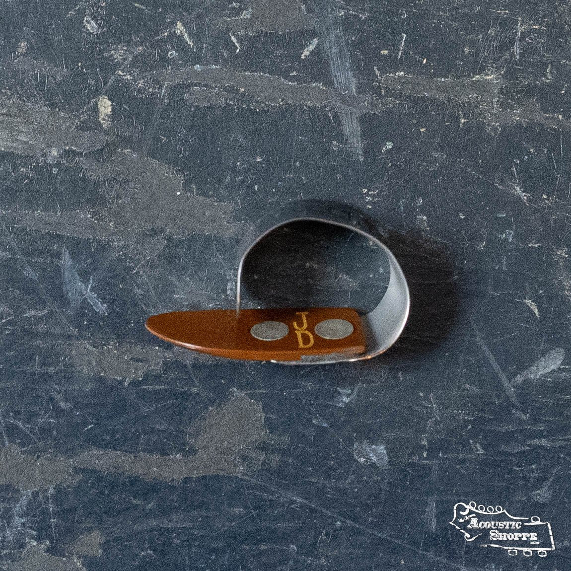 A BlueChip Picks (BCT-JDS) thumb pick by BlueChip with a brown plastic tip rests on a scratched, dark gray surface. The Acoustic Shoppe logo appears in the bottom right corner.