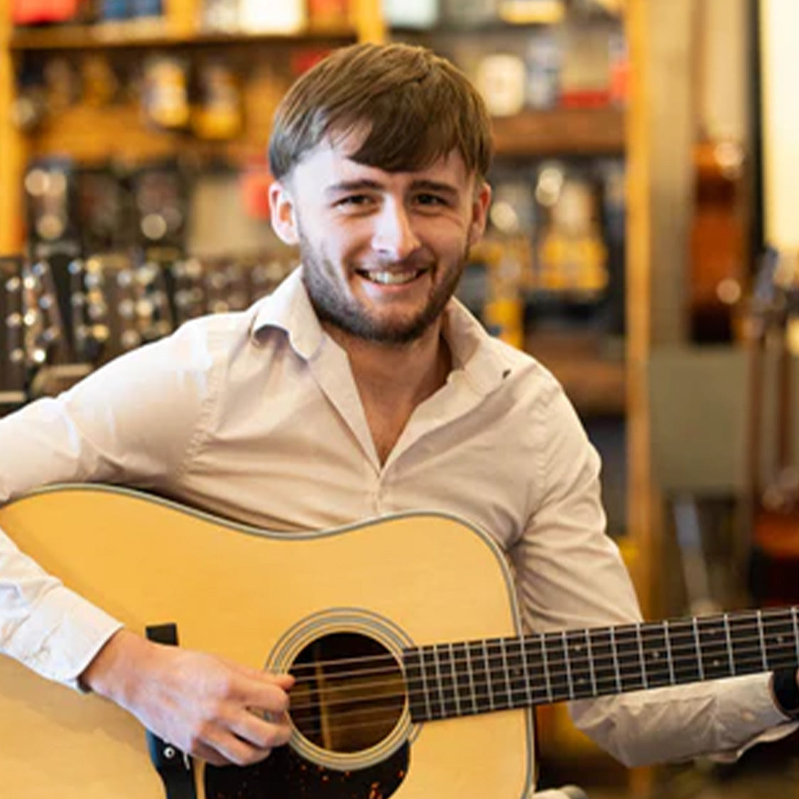 A young man with short brown hair and a beard smiles while playing The Acoustic Shoppe’s *New 2025 Model* Martin DE Retro Plus Torrefied Spruce/Mahogany Dreadnought Guitar w/Martin Electronics indoors, guitars and shelves behind him.