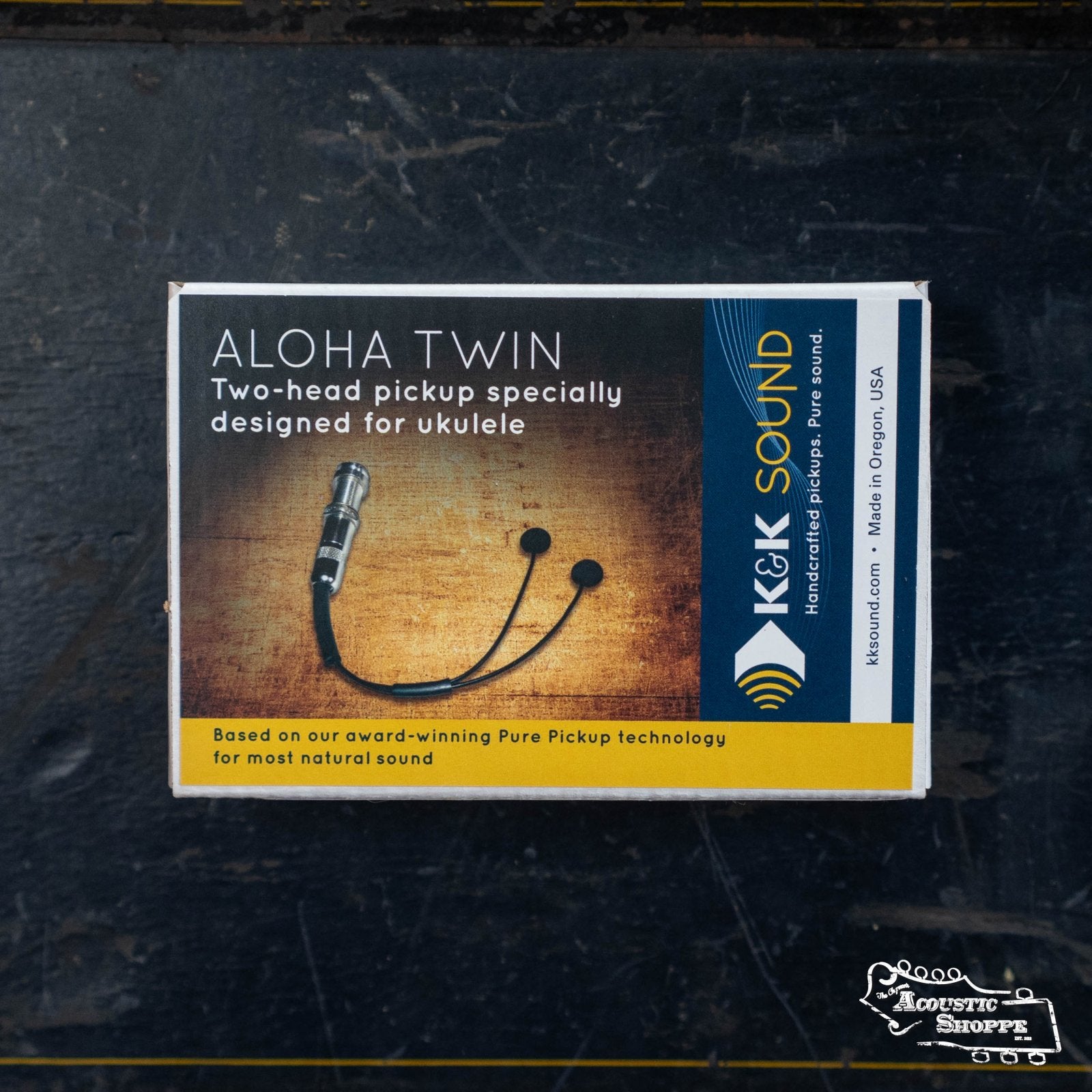 A box labeled K&K Aloha Twin Ukulele Pickup by K&K Sound displays a two-head bridge plate pickup, featuring an image of the product, company info, and The Acoustic Shoppe logo on a dark background.