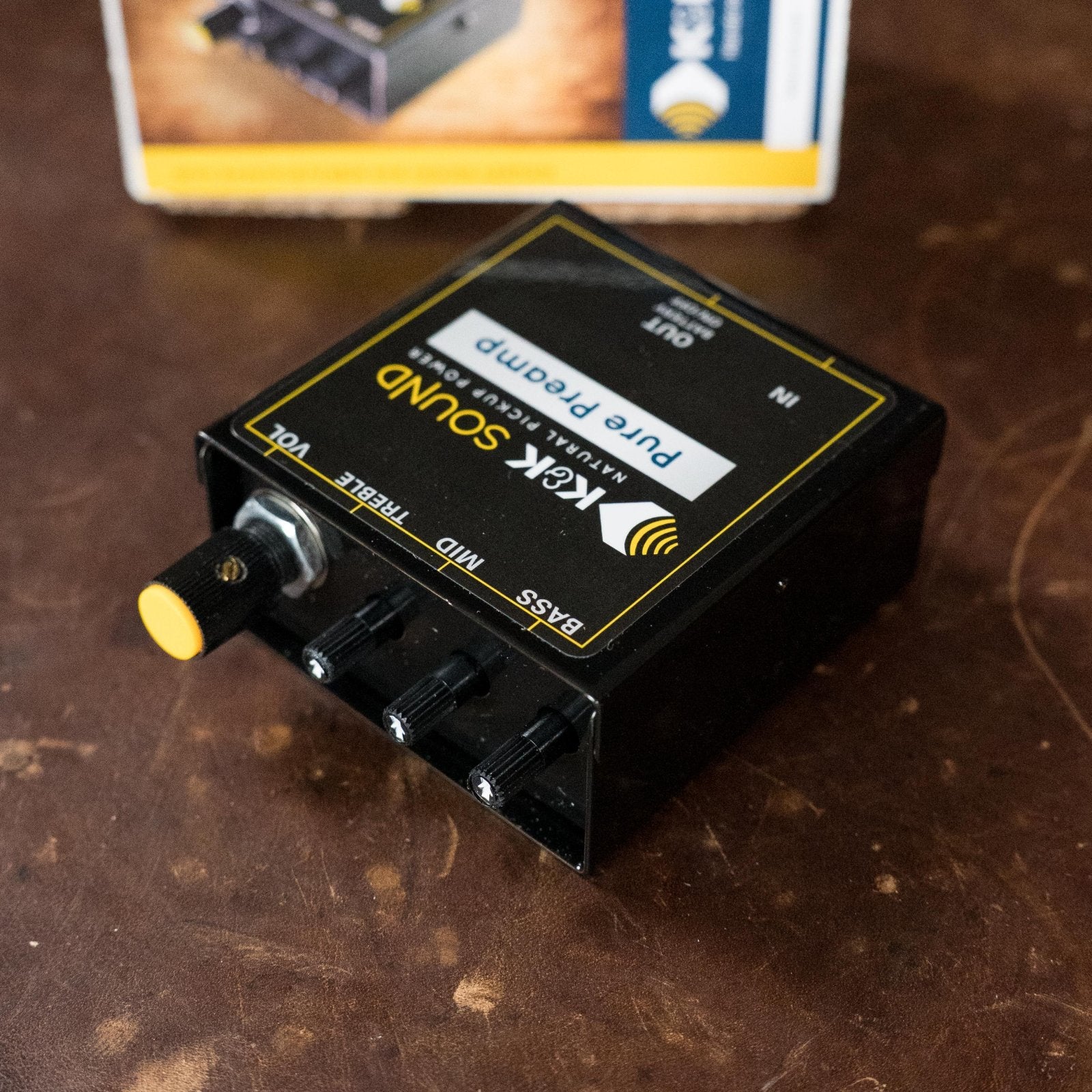 A black K&K Sound Pure Preamp with yellow and white labeling sits on a brown surface, featuring volume, bass, mid, treble knobs, and an internal gain trimmer. Part of its box appears in the background.