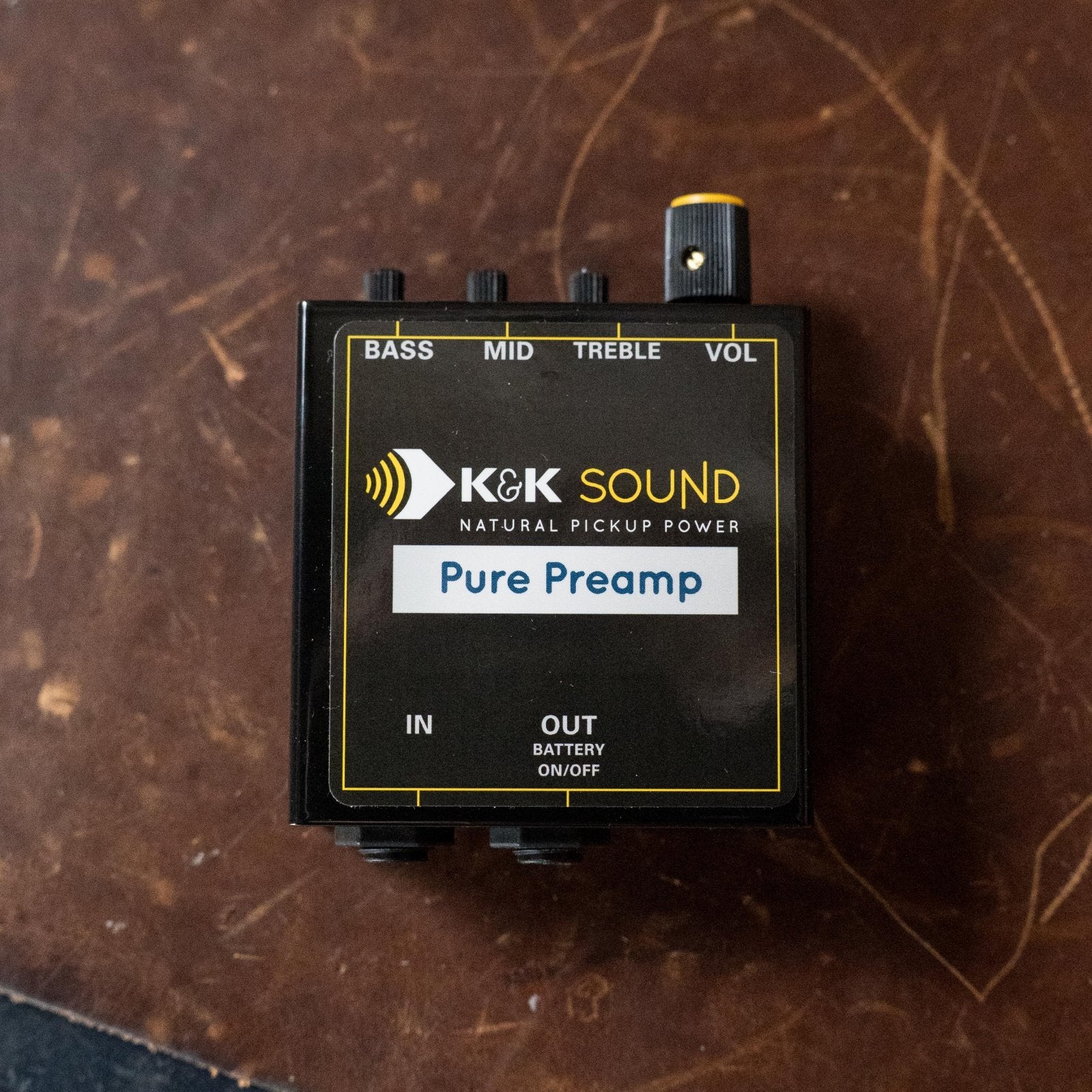 A K&K Sound K&K Pure Preamp with bass, mid, treble, and volume controls plus an internal gain trimmer rests on brown leather. It includes input/output jacks and a battery indicator switch.