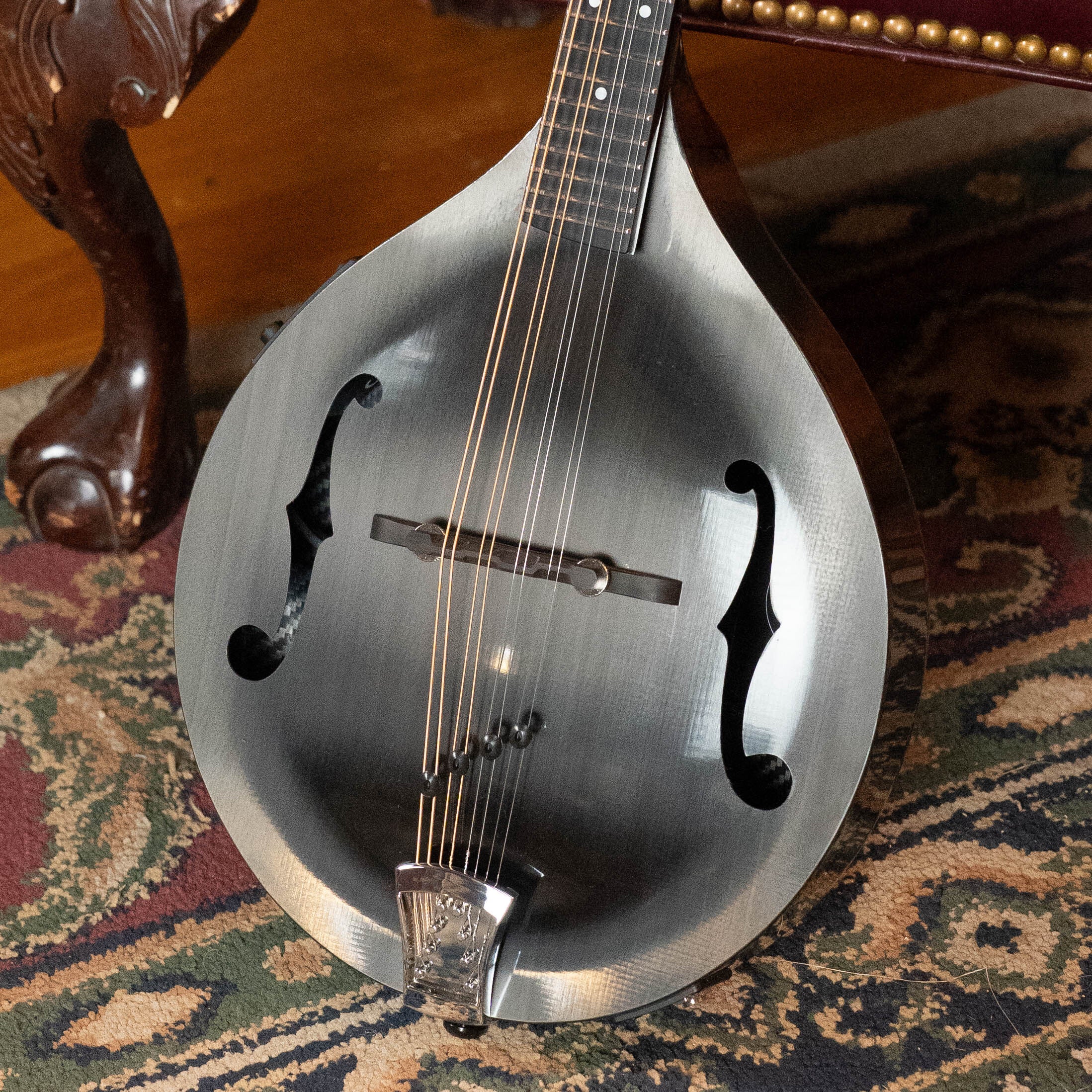 A close-up of the KLOS Guitars CT_MA_DAE_EQ Full Carbon Unidirectional Black A-Style Mandolin with KLOS EQ Pickup (#4889), featuring f-shaped sound holes, resting on a patterned rug near a carved wooden chair.