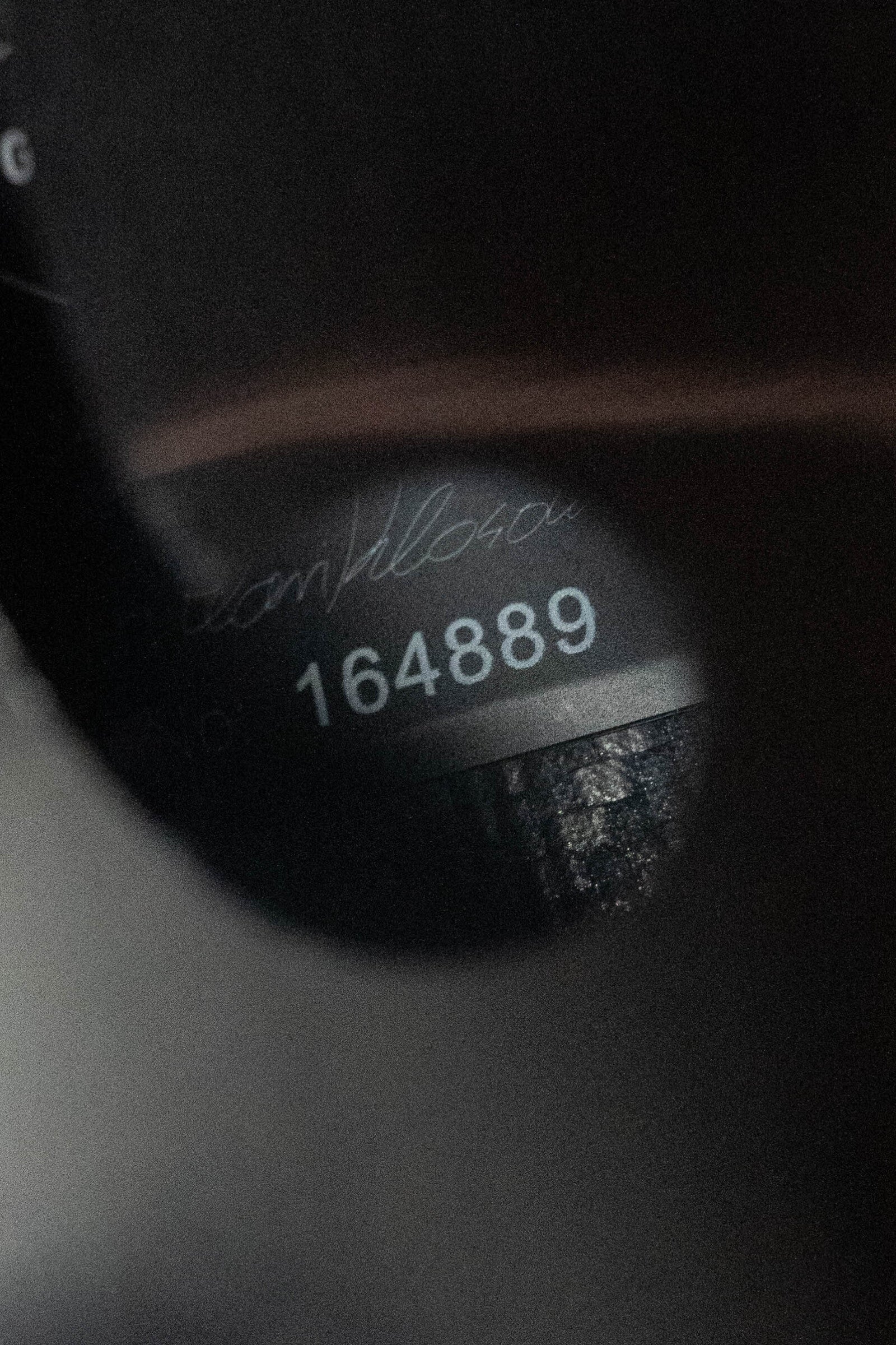 Close-up of a KLOS Guitars CT_MA_DAE_EQ Full Carbon A-Style Mandolin (#4889), showing the number 4889 through an opening, with a blurred background and faint handwritten text above the number.