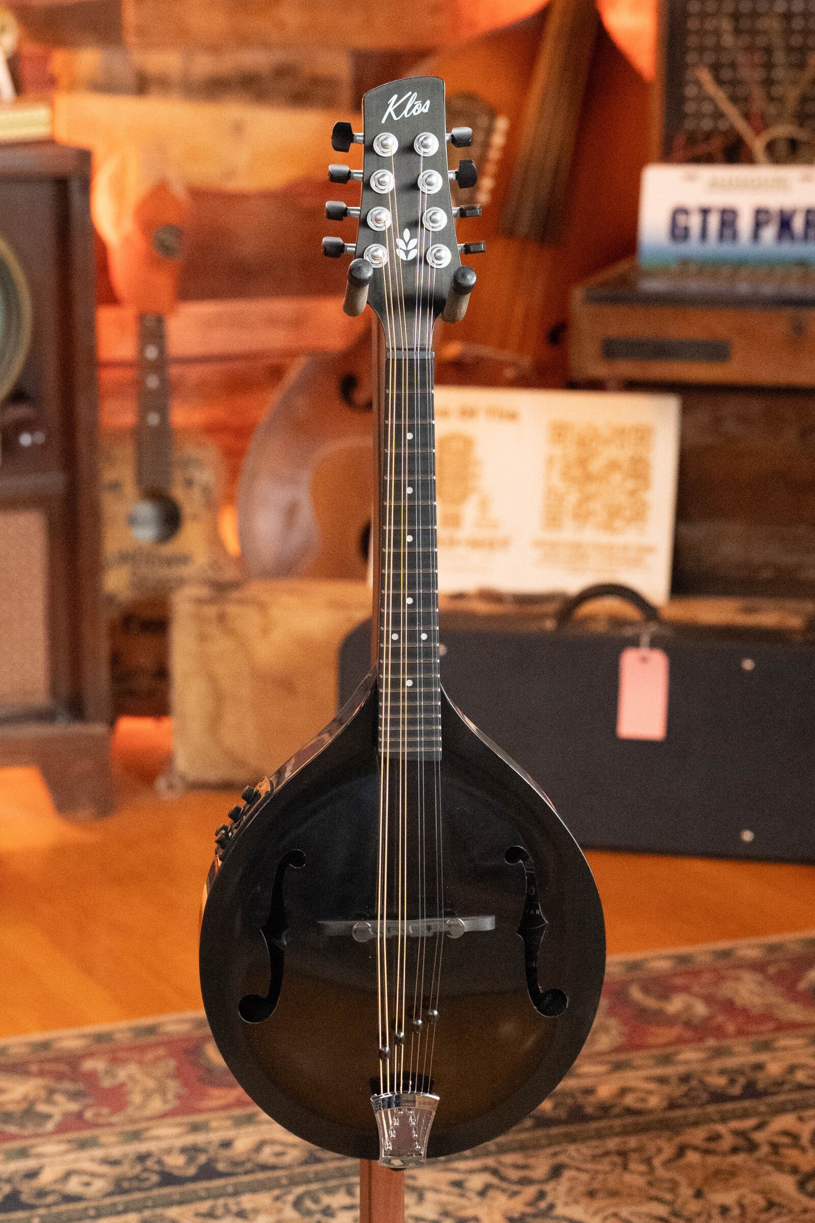 A KLOS Guitars CT_MA_DAE_EQ Full Carbon Unidirectional Black A-Style Mandolin w/KLOS EQ Pickup (#4889) with eight strings and f-holes sits on a stand in a cozy, wood-paneled room among musical instruments and equipment.