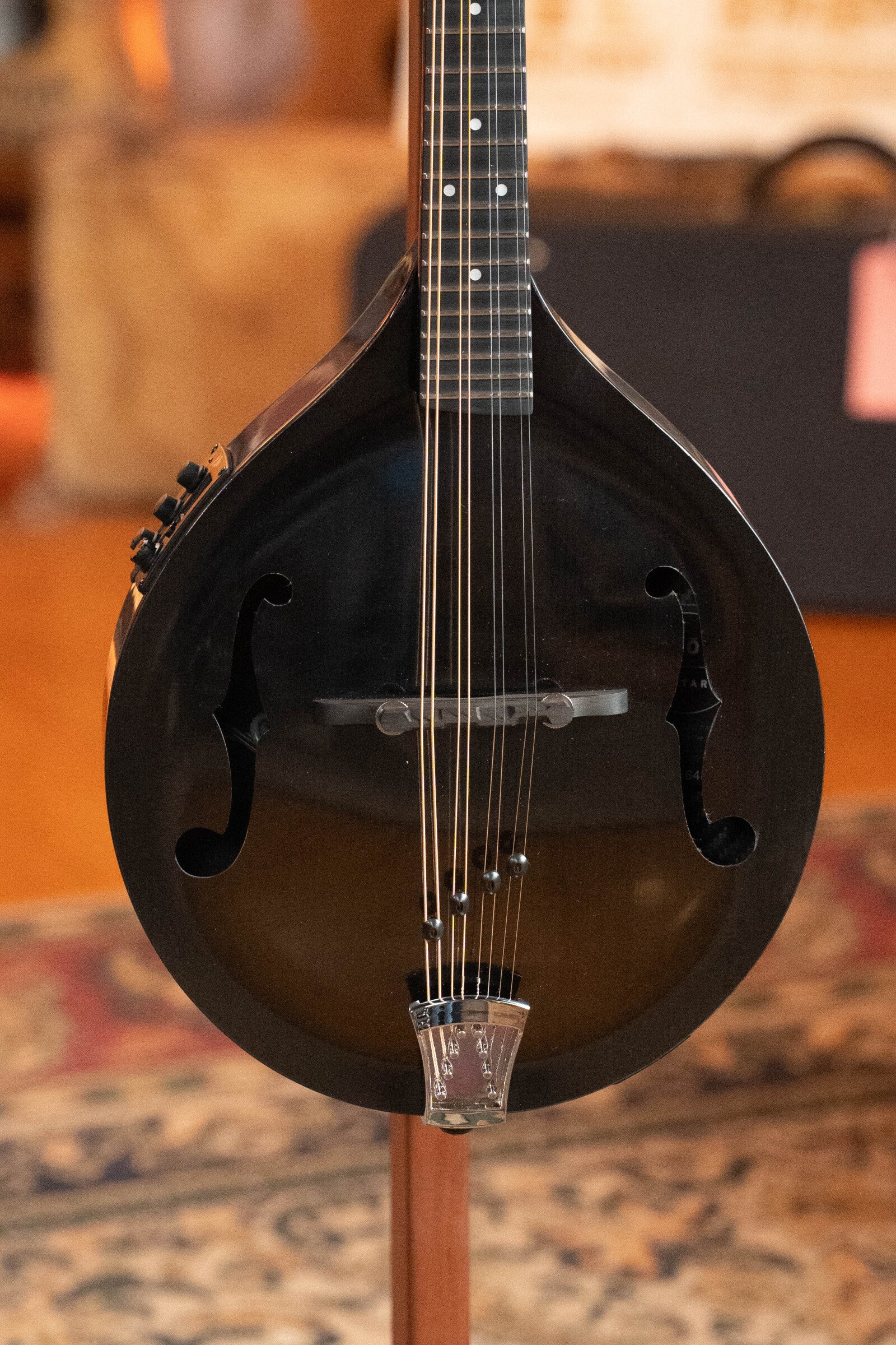 A close-up of the KLOS Guitars CT_MA_DAE_EQ Full Carbon Unidirectional Black A-Style Mandolin with KLOS EQ Pickup #4889 showcases its body, strings, and f-shaped sound holes against a patterned rug and blurred background.