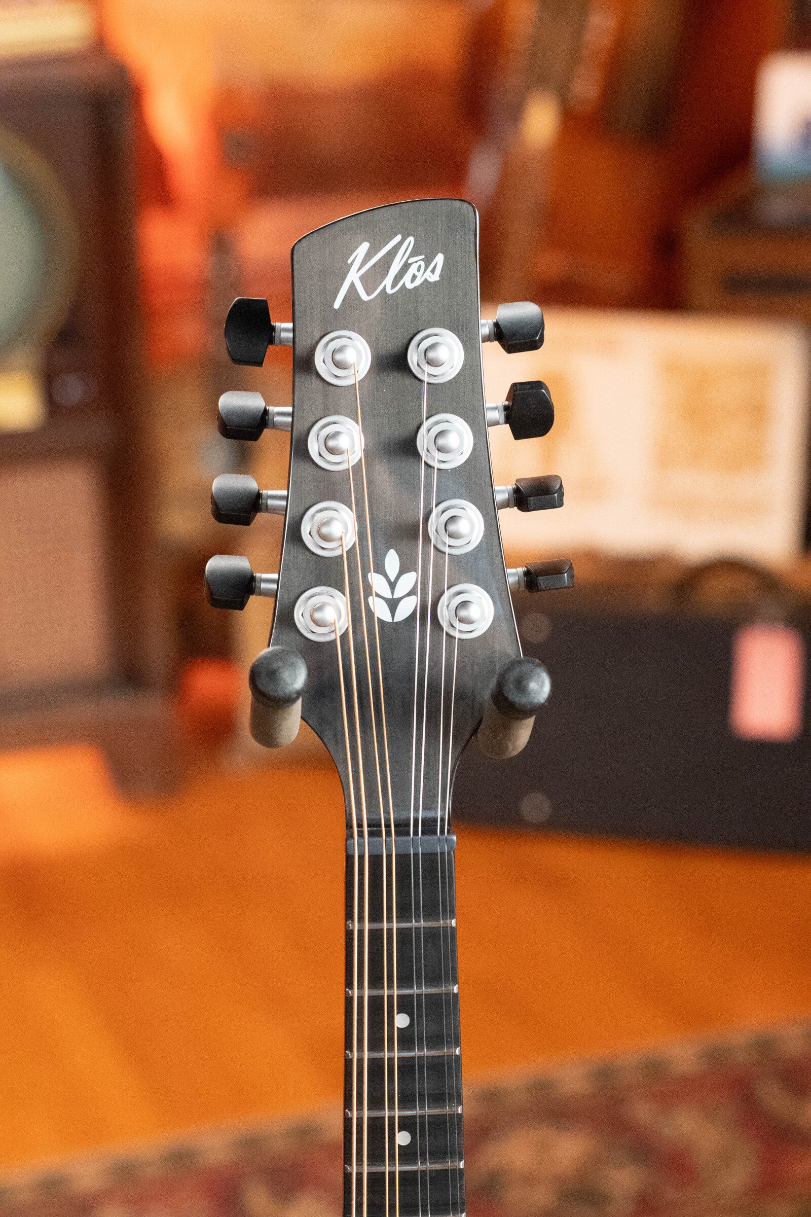 Close-up of a KLOS Guitars CT_MA_DAE_EQ Full Carbon A-Style Mandolin (#4889) headstock with eight tuning pegs and a white leaf design, set against a warm, blurred background with wooden tones and musical gear.