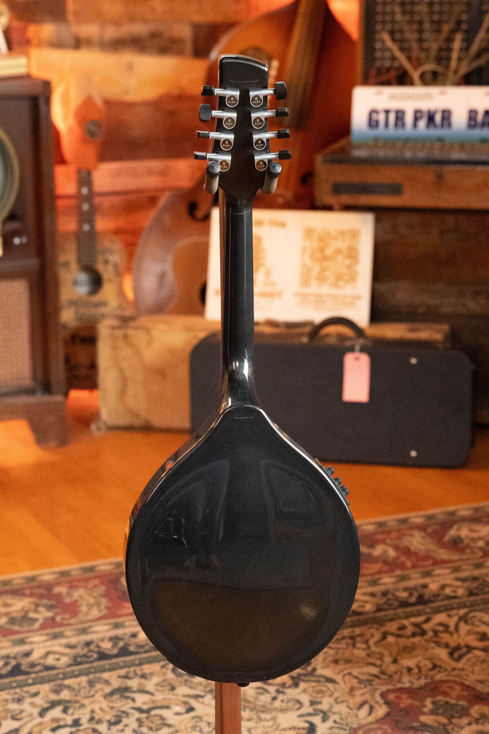 A KLOS Guitars CT_MA_DAE_EQ Full Carbon Unidirectional Black A-Style Mandolin (#4889) with KLOS EQ Pickup is shown from the back on a stand in a cozy, wood-paneled room with vintage decor and patterned carpet.