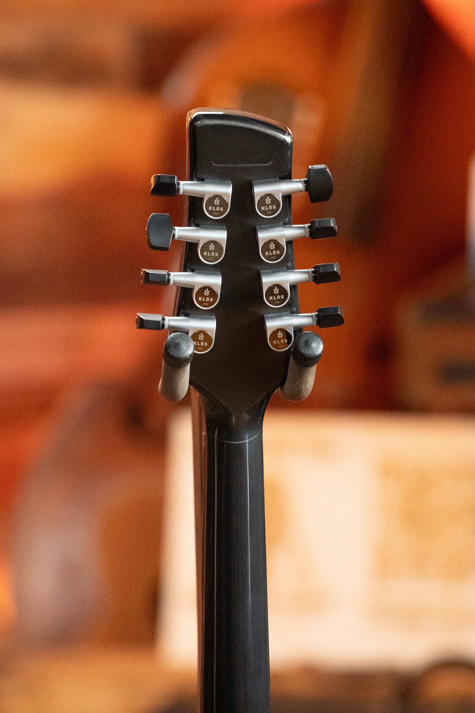 Close-up of the KLOS Guitars CT_MA_DAE_EQ Full Carbon Unidirectional Black A-Style Mandolin (#4889) headstock, showing six tuning pegs and part of the neck against a warm, blurred background.