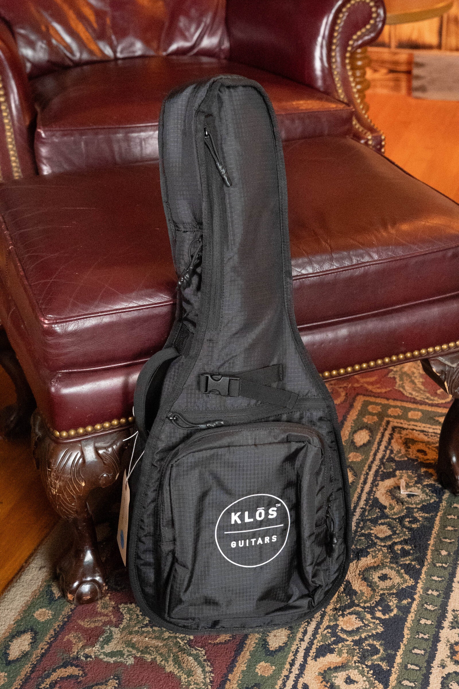 A KLOS Guitars CT_MA_DAE_EQ Full Carbon Unidirectional Black A-Style Mandolin with KLOS EQ Pickup (#4889) in its black gig bag stands upright on a patterned rug before a maroon leather chair and ottoman indoors.