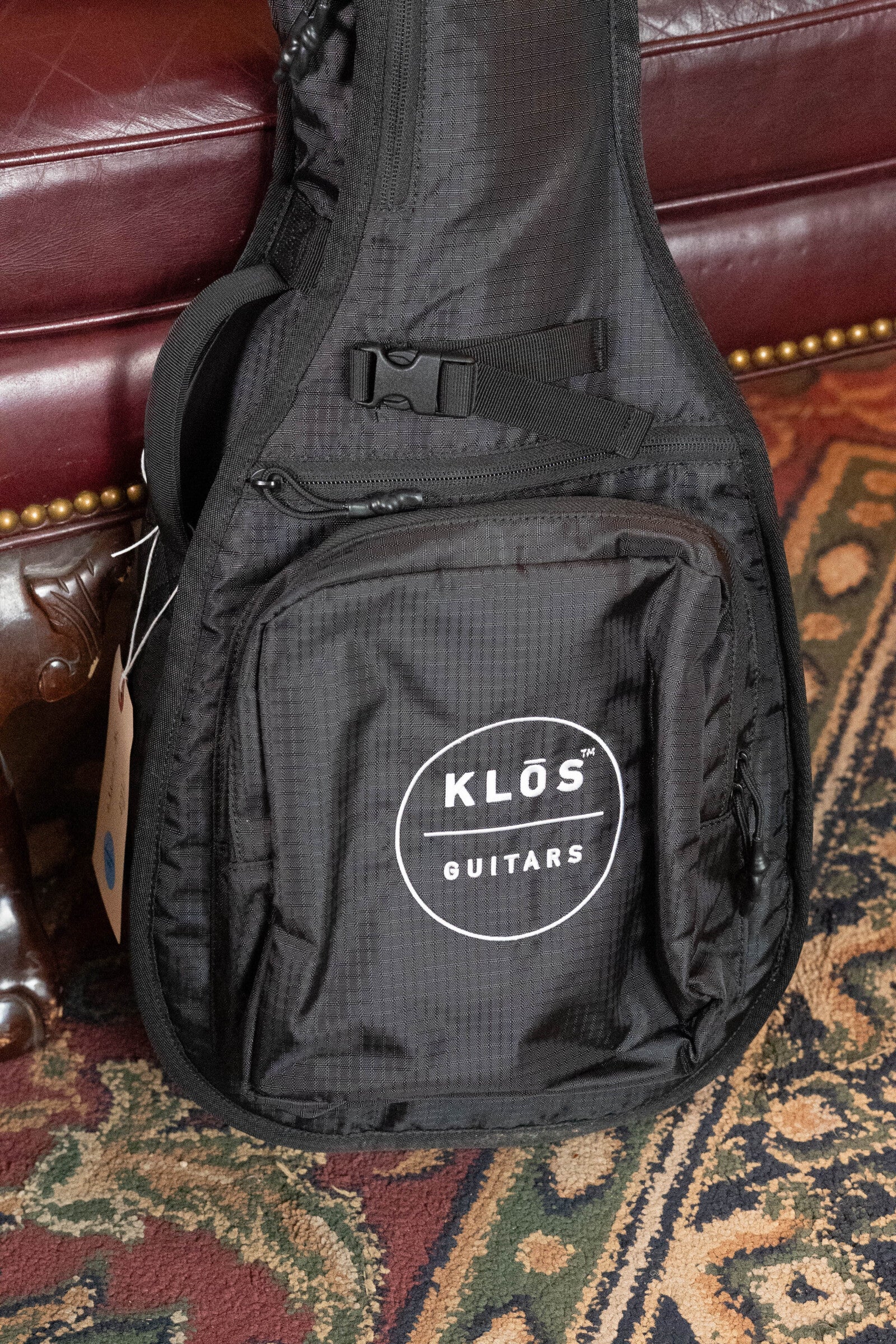 A KLOS Guitars CT_MA_DAE_EQ Full Carbon Unidirectional Black A-Style Mandolin w/KLOS EQ Pickup (#4889) sits on a patterned rug beside a brown leather couch with brass studs.