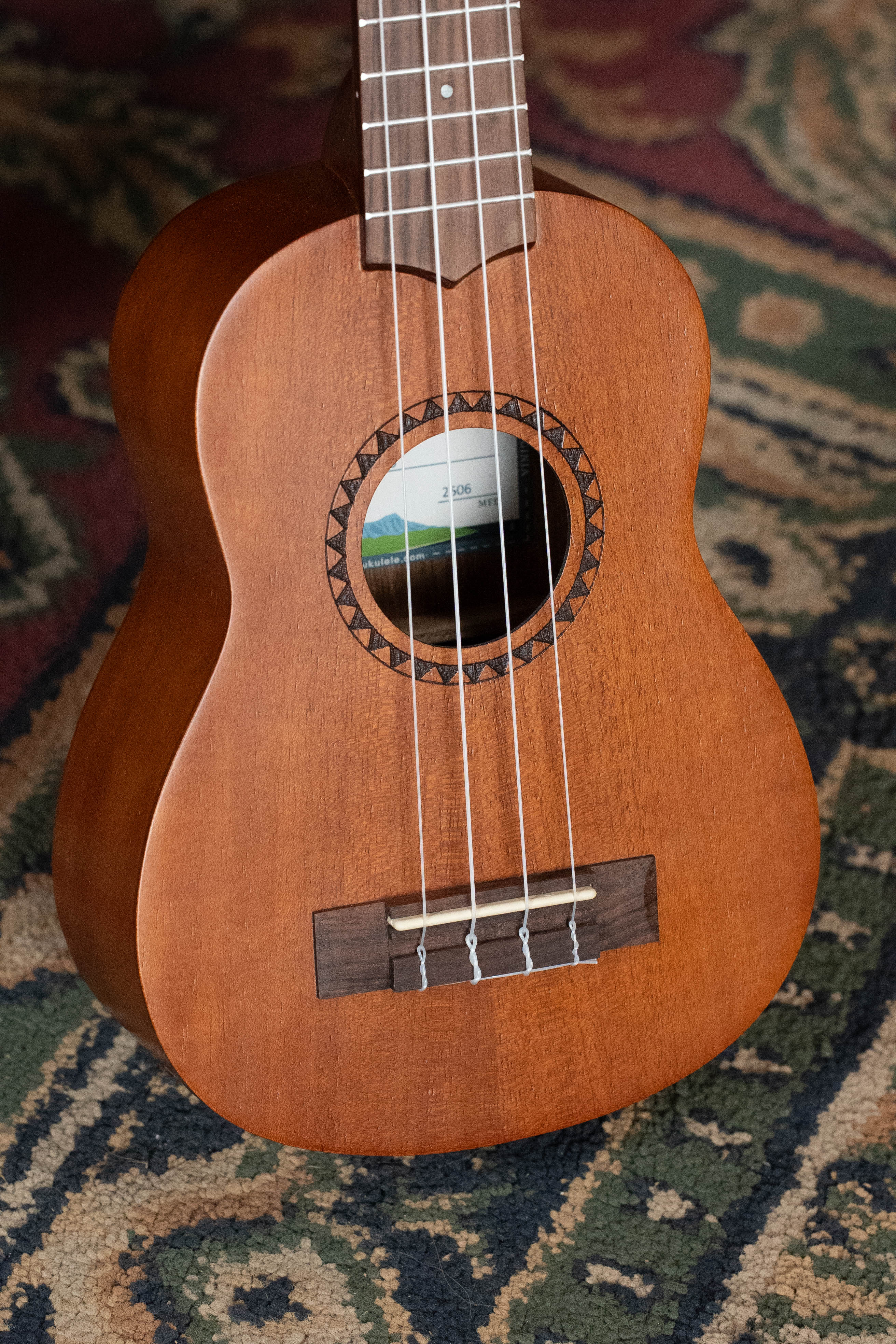 Soprano ukulele with satin mahogany body and wooden bridge on patterned rug background. - Kala KA-15S Satin Mahogany Soprano Ukulele - Default Title