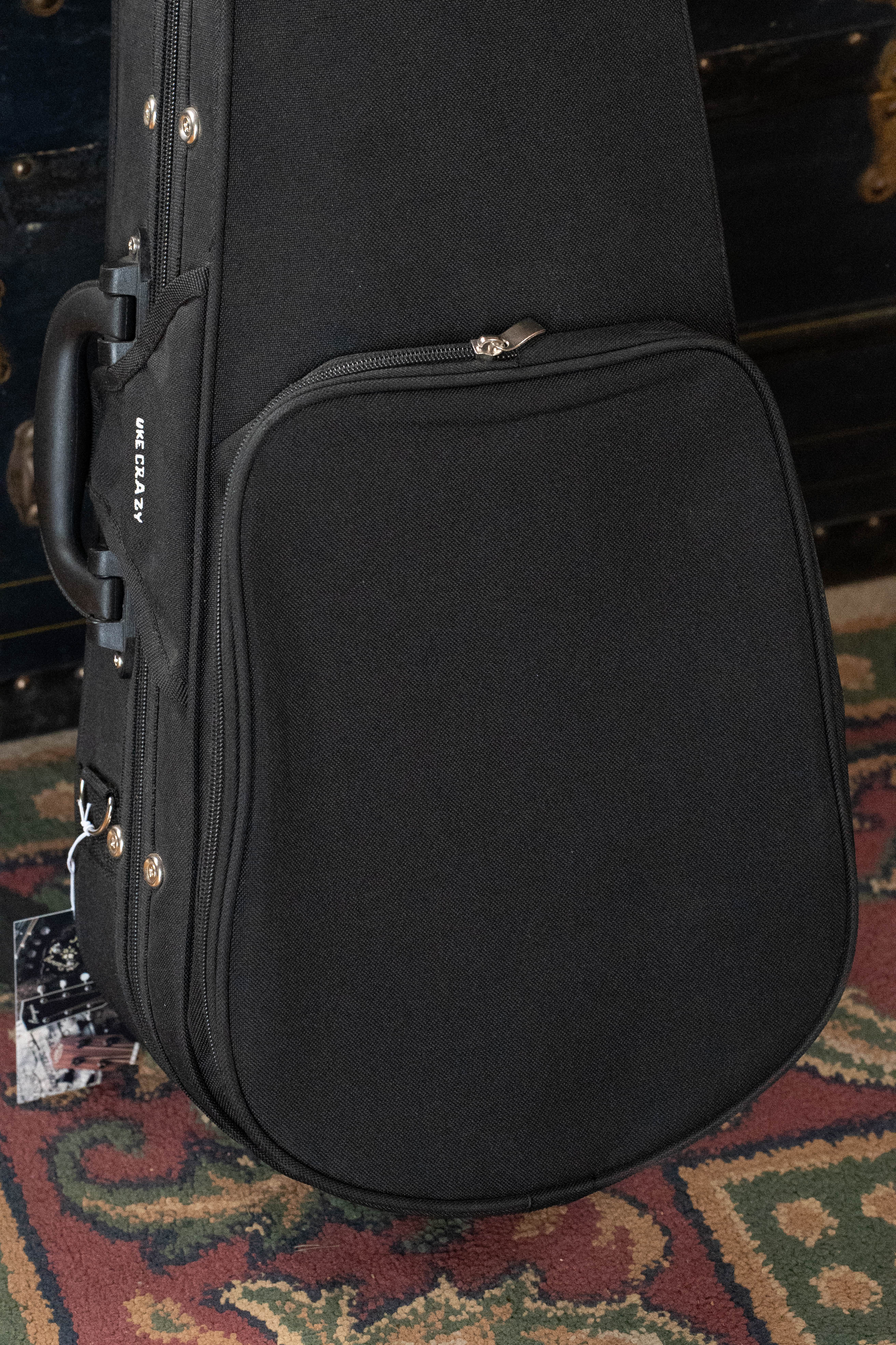 A black Kala UC-C Foam Concert Ukulele Hardcase with a large front pocket and handle stands upright on a patterned rug. The Kala brand name is visible on the side of the case.