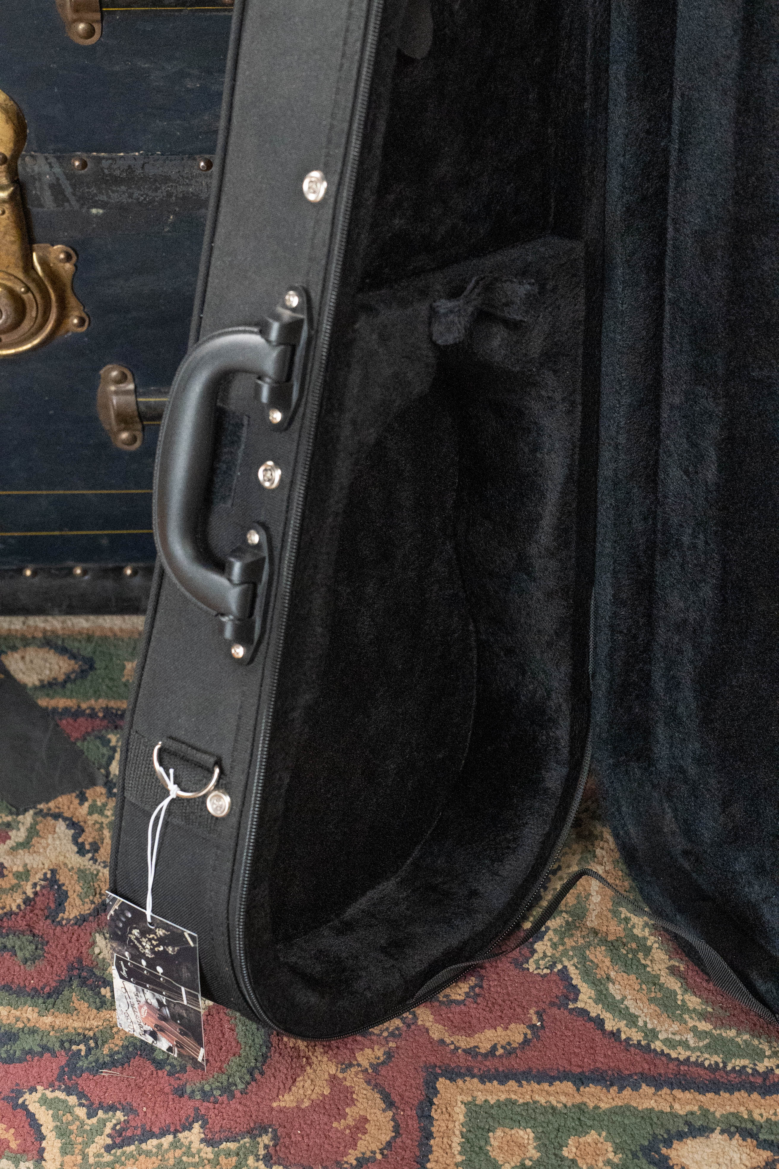 A close-up of an open, empty Kala UC-C Foam Concert Ukulele Hardcase, lined with soft material, resting on a patterned rug beside a dark trunk with brass fittings. A tag is attached to the Kala concert ukulele case handle.