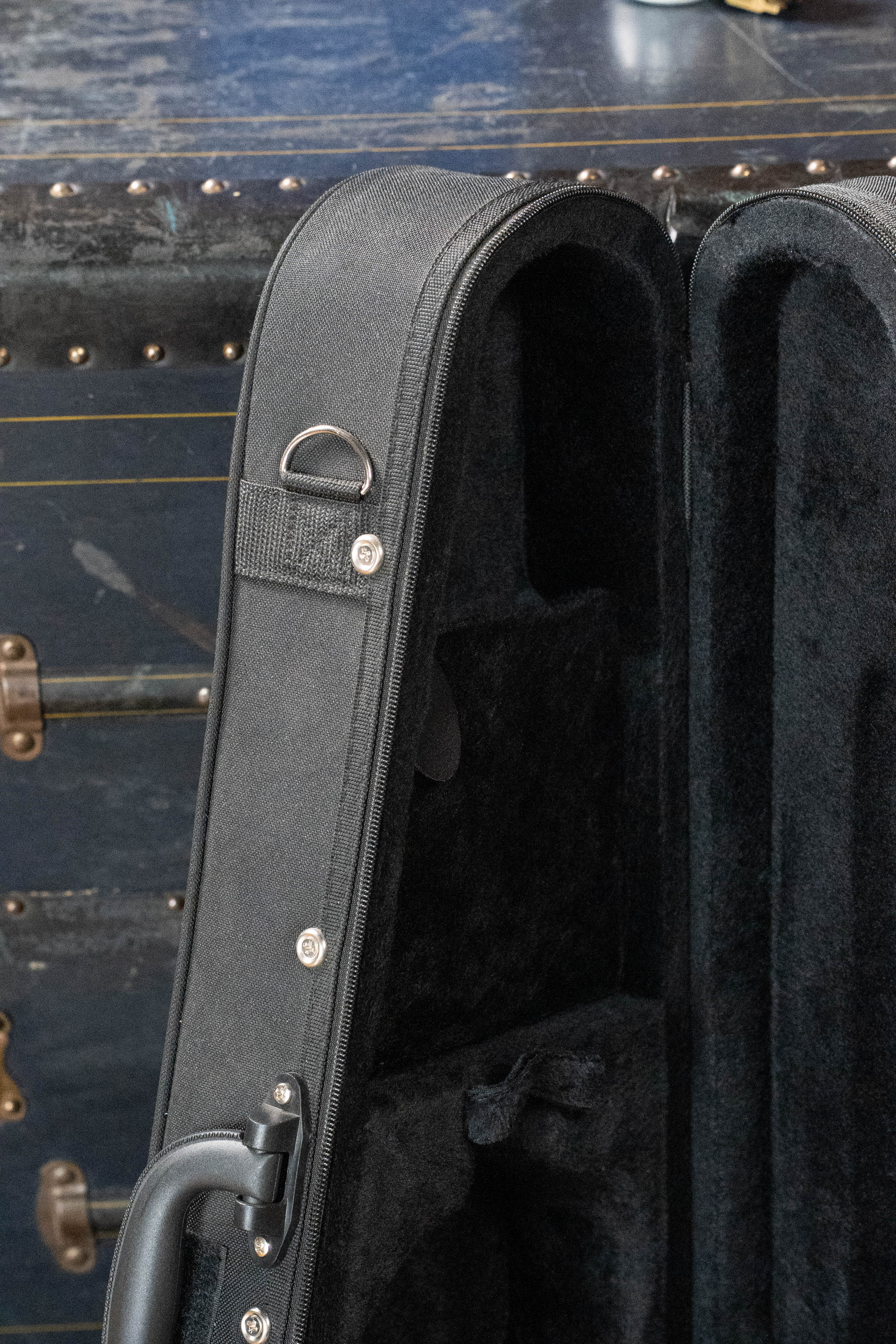 A close-up of an open Kala UC-C Foam Concert Ukulele Hardcase by Kala, featuring a black velvet lining, set against a dark, textured trunk with metal rivets and latches in the background.