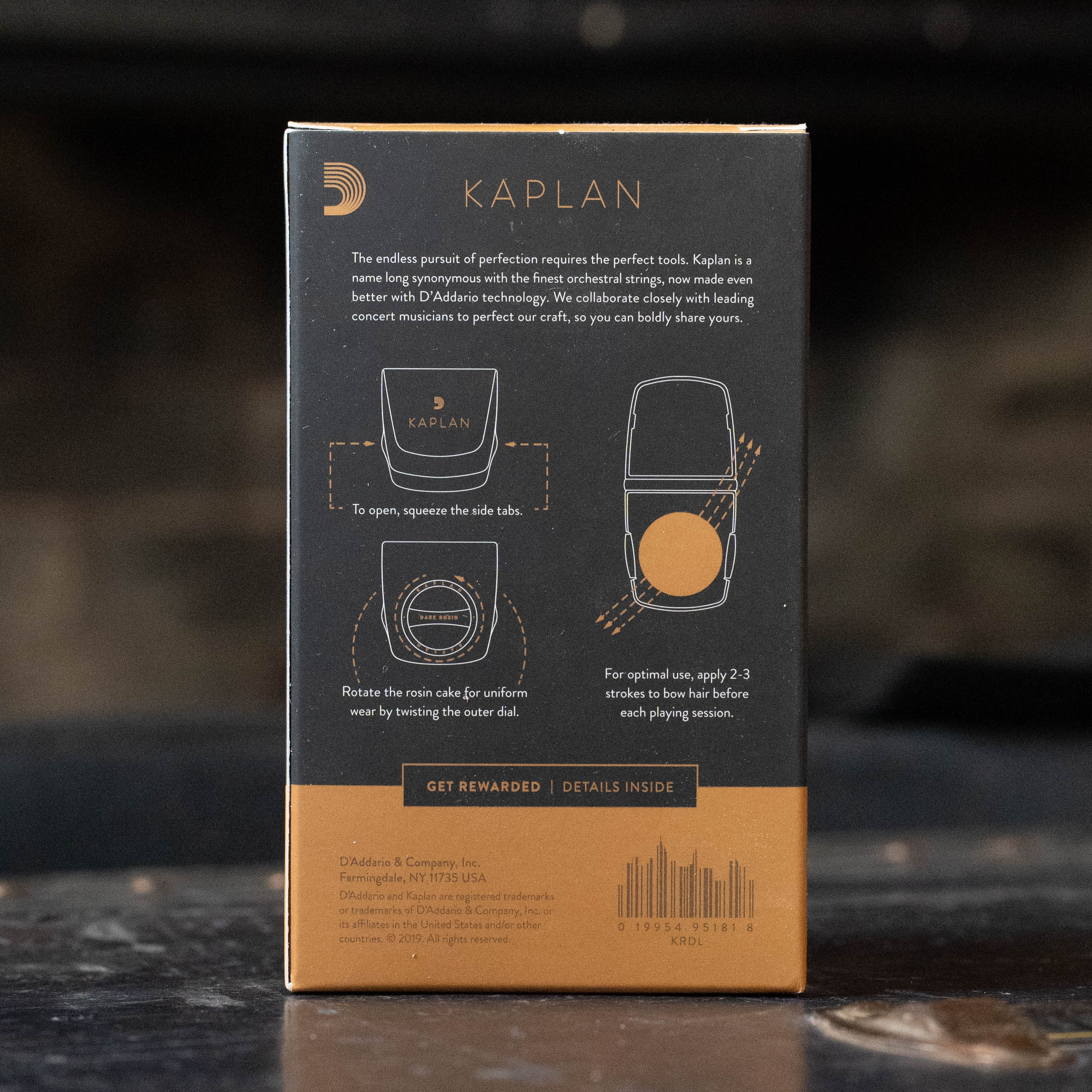 A box labeled D'Addario Kaplan Premium Rosin with Case (KRD) displays illustrated opening and usage instructions for the low-dust violin rosin, plus text and a barcode at the bottom. The background appears out of focus.