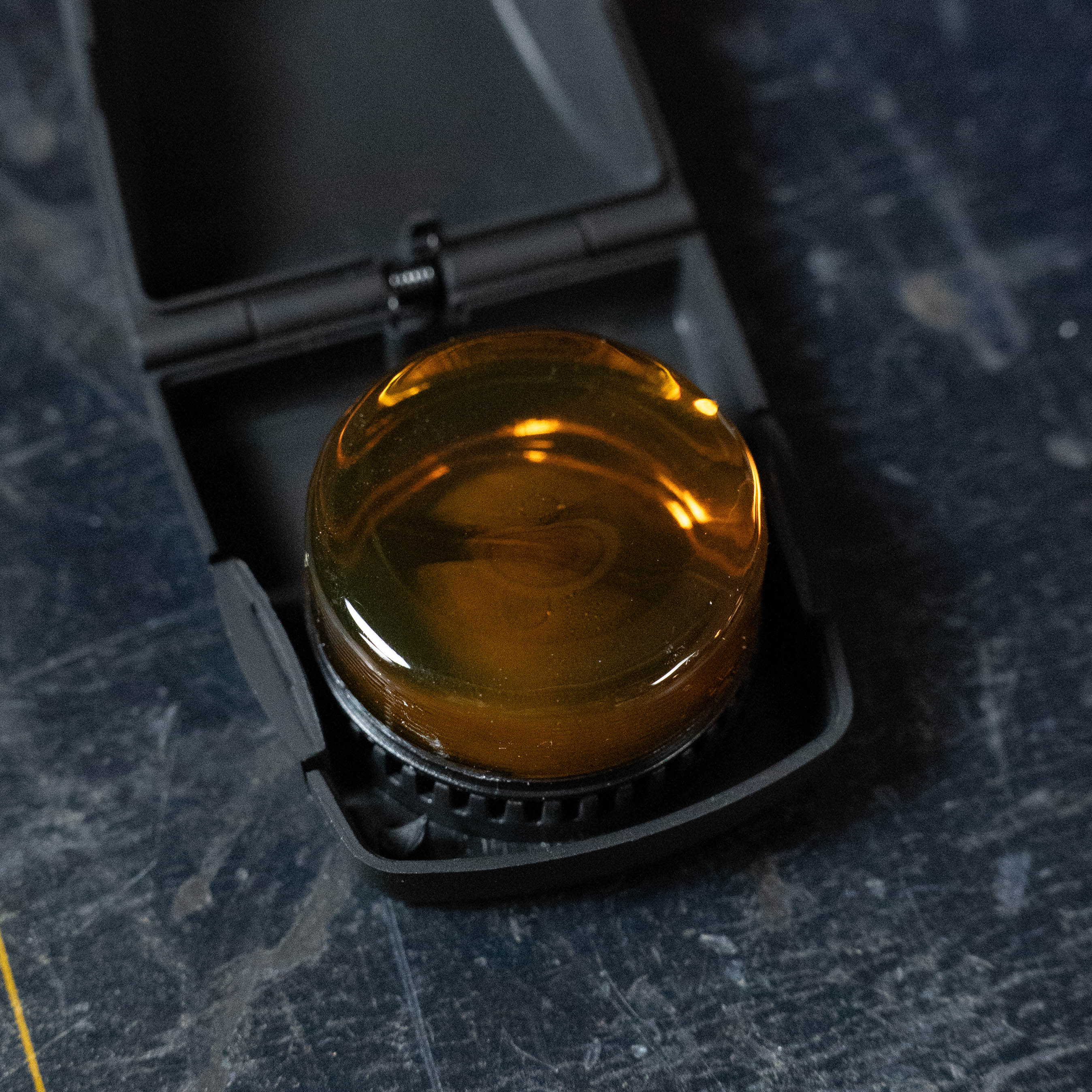A round, amber D'Addario Kaplan Premium Rosin with Case (KRD) by Kaplan rests in its open black protective case on a dark, textured surface, showing its shiny, low-dust surface ideal for violinists.