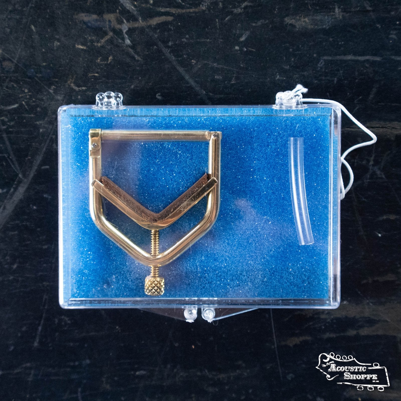 The Kat Eyz Brass 0 Radius Banjo Capo (1/4 TS) by Kat Eyz rests on blue foam in a clear, hinged plastic box. Included are a short clear tube and white string. The case is placed on a dark, textured surface.