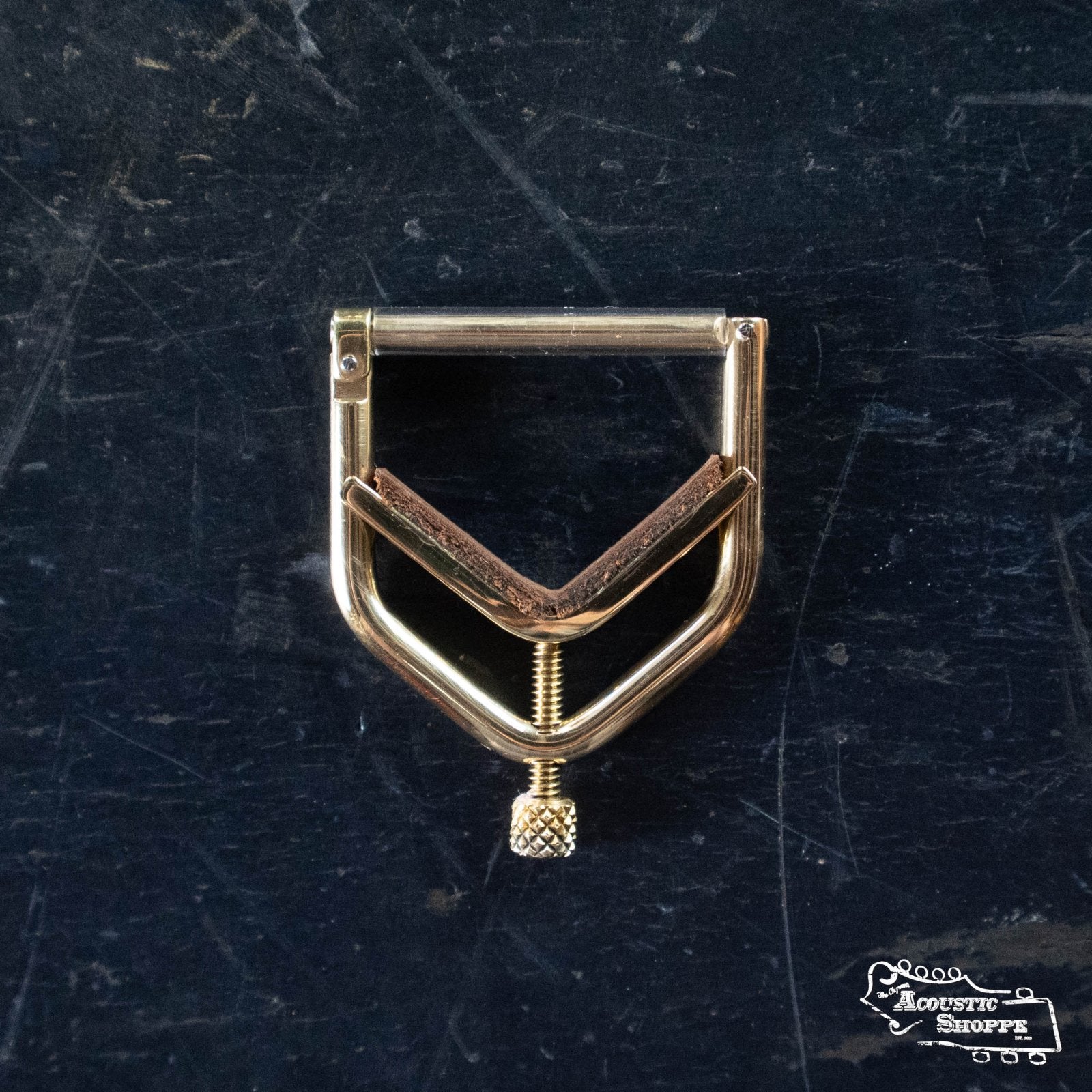 A Kat Eyz Brass 0 Radius Banjo Capo (1/4 TS) with a screw tightening mechanism and leather padding sits on a dark, scratched surface. “The Acoustic Shoppe” logo is visible in the bottom right corner.