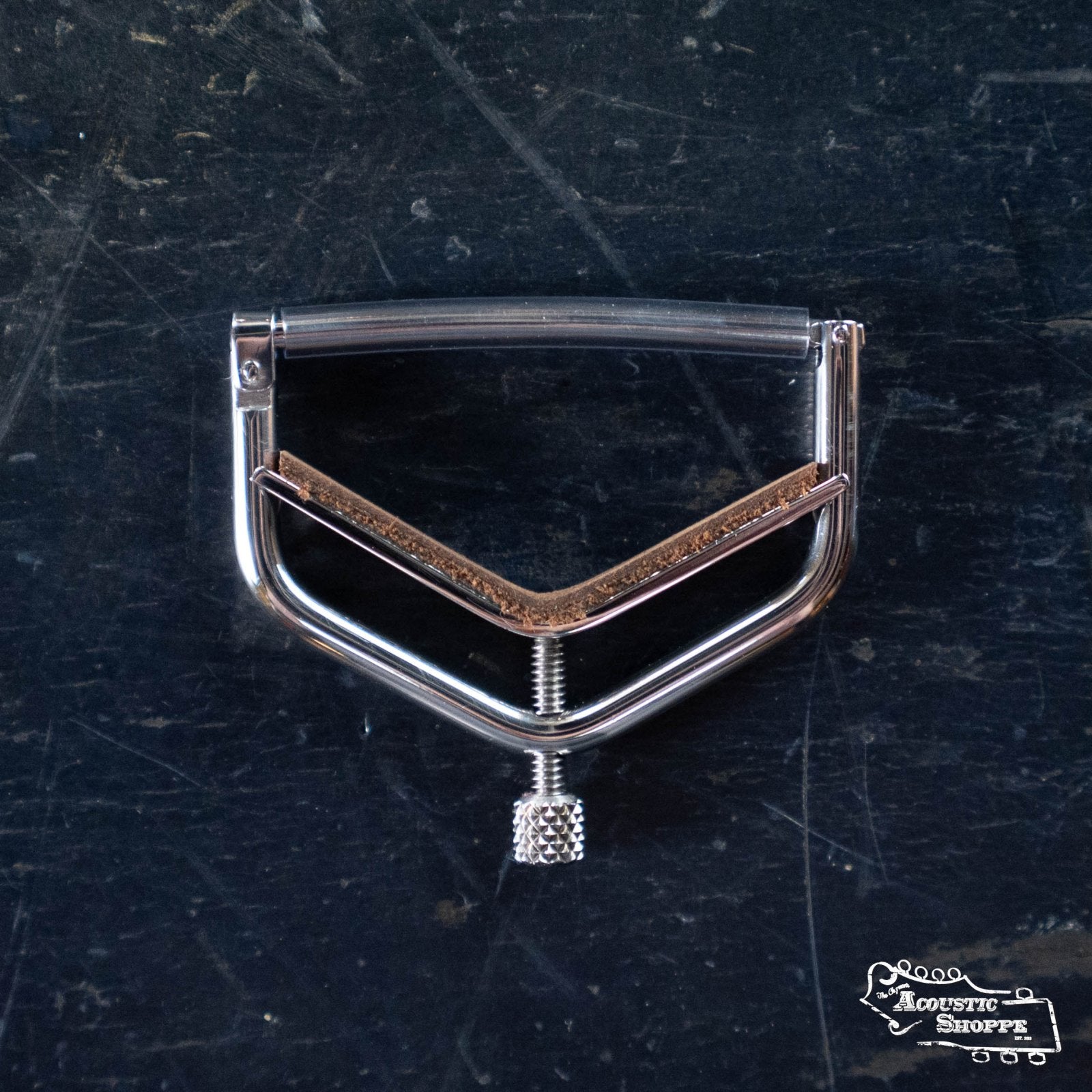 The Kat Eyz Stainless Steel 12 Radius Guitar Capo (1/4 TS), a handmade capo with a V-shaped design and cork padding, rests on a scratched dark surface. It features a screw adjustment and the Acoustic Shoppe logo in the lower right corner.