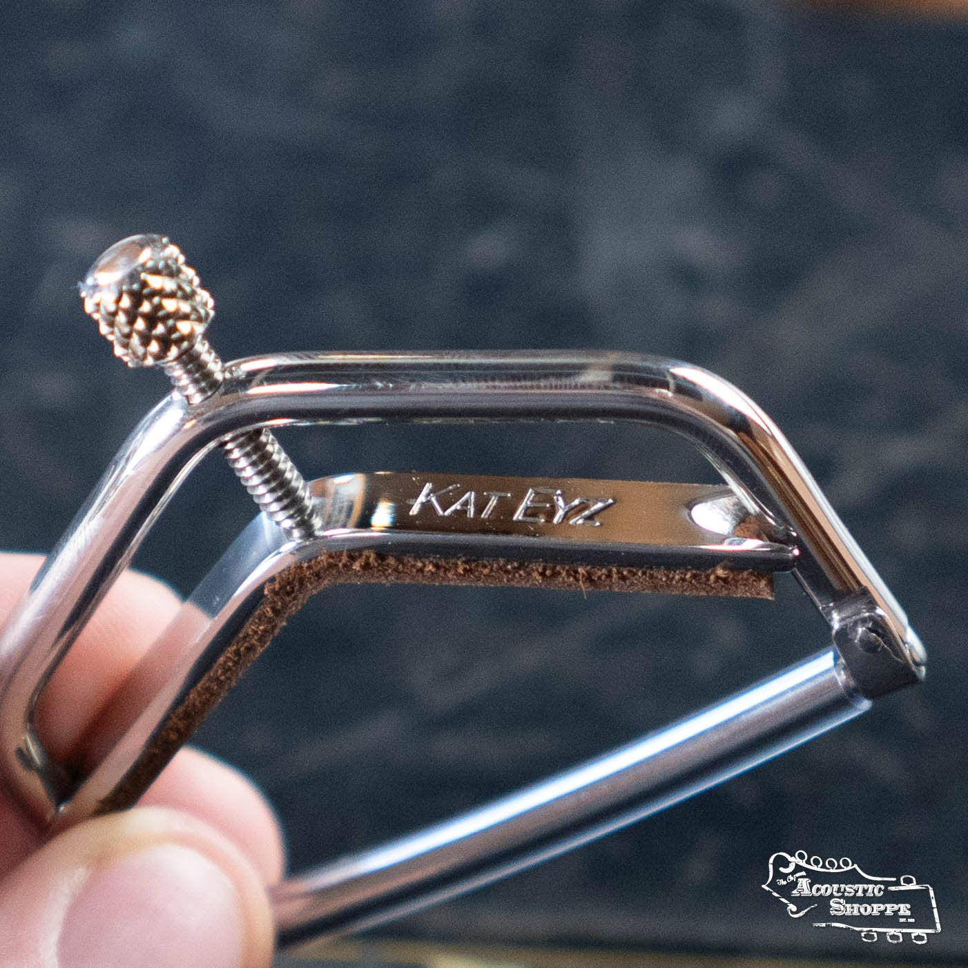A close-up of a hand holding the Kat Eyz Stainless Steel 12 Radius Guitar Capo (1/4 TS), showing its textured adjustment screw and leather pad. The Acoustic Shoppe logo appears in the bottom right corner.