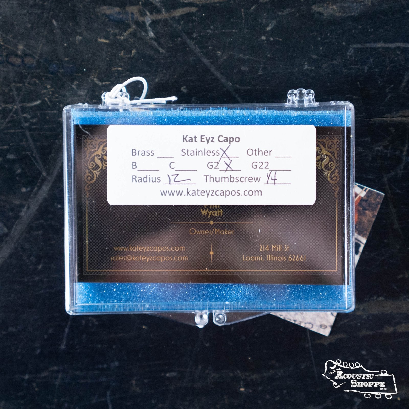 A blue glittery case holds a Kat Eyz Stainless Steel 12 Radius Guitar Capo (1/4 TS) card, listing handmade capo options and contact info, set on a dark surface.
