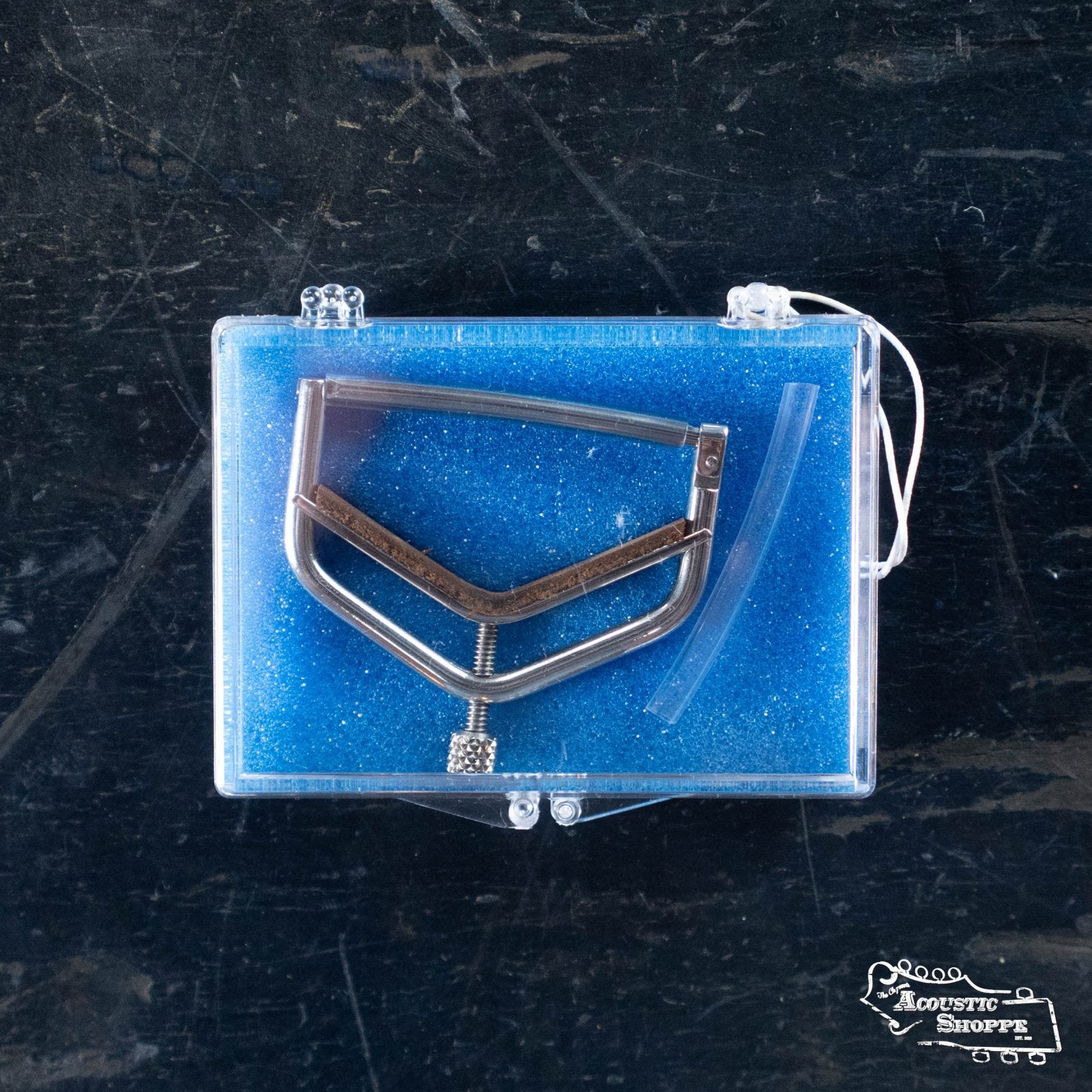 A Kat Eyz Stainless Steel 16 Radius Guitar Capo (1/4 TS) is displayed on blue foam in a clear case, resting on a dark surface. The Acoustic Shoppe logo appears at the bottom right of this premium handmade capo presentation.