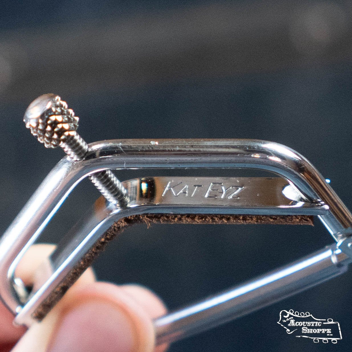 Close-up of a hand holding a Kat Eyz Stainless Steel 16 Radius Guitar Capo (1/4 TS), engraved with KAT EYZ and featuring screw adjustment. The Acoustic Shoppe logo appears in the lower right corner.