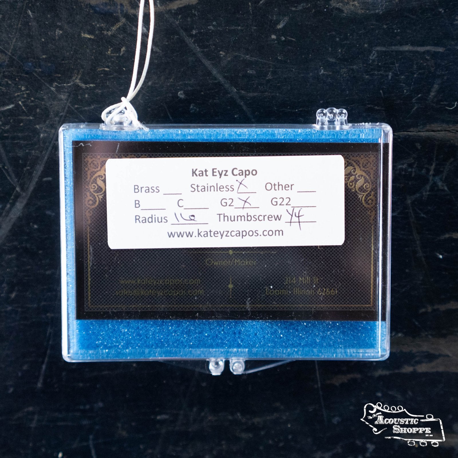 A blue glittery box labeled Kat Eyz displays the Kat Eyz Stainless Steel 16 Radius Guitar Capo (1/4 TS), with handwritten marks on Stainless, C, and Thumbscrew—showcasing this handmade capo. Website info appears below.