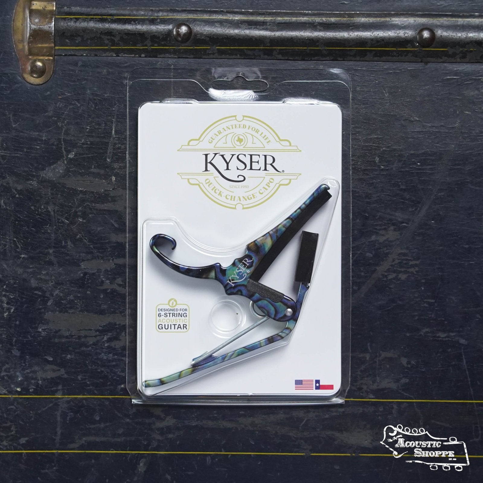 A Kyser 6 String Quick Change Abalone Capo, featuring a vibrant marbled design, is shown in its packaging on a dark textured surface with the Acoustic Shoppe logo in the bottom right corner.
