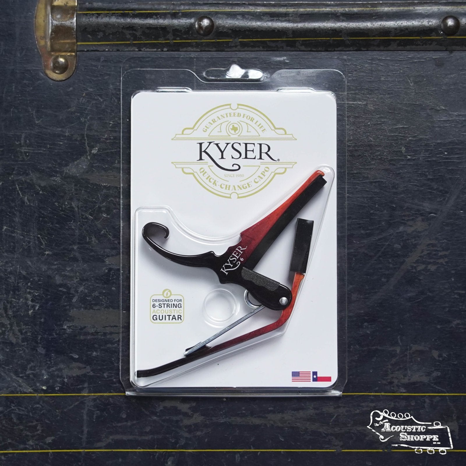 A Kyser 6 String Quick Change Sunburst Capo is shown in its packaging on a dark surface. The packaging features the Kyser brand and a small American flag. This spring-loaded capo comes in a black and red sunburst finish.