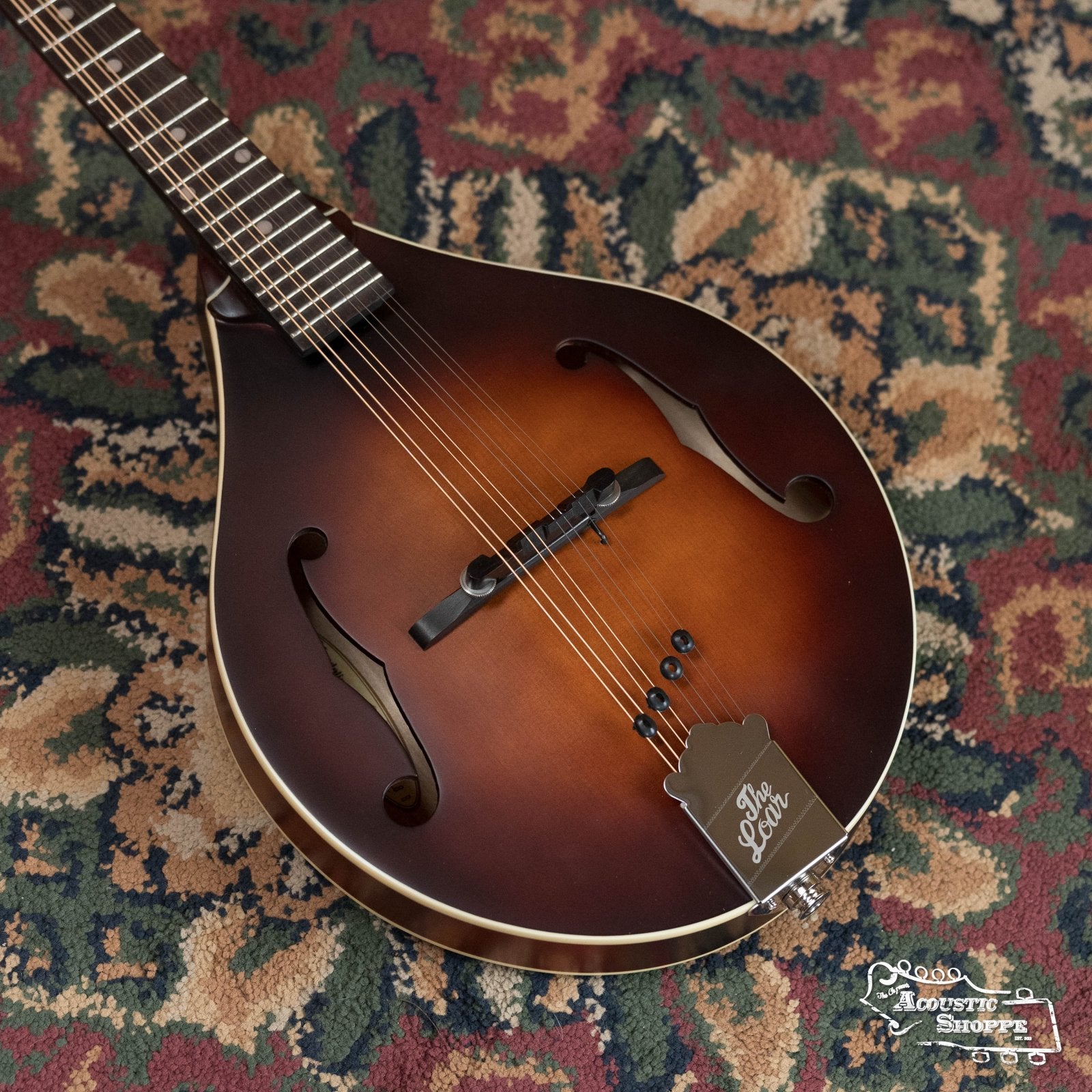 A The Loar LM-110E-BRB "Honey Creek" A-Style Brownburst Mandolin with f-shaped sound holes and a solid spruce top lies on a patterned carpet. The metal tailpiece reads The Loar, with the Road Acoustic Shoppe logo in the lower right corner.