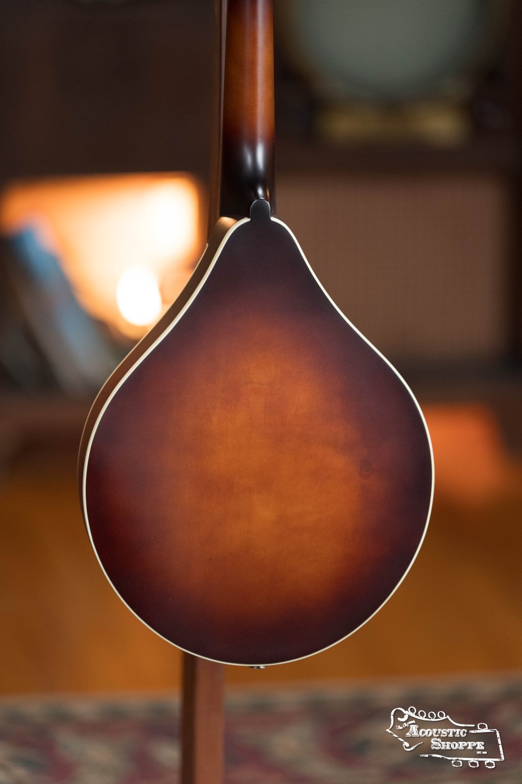 A close-up of the back of The Loar LM-110E-BRB "Honey Creek" A-Style Brownburst Mandolin w/Pickup #0688 stands upright indoors on a patterned rug, with warm lighting and furniture softly blurred in the background.