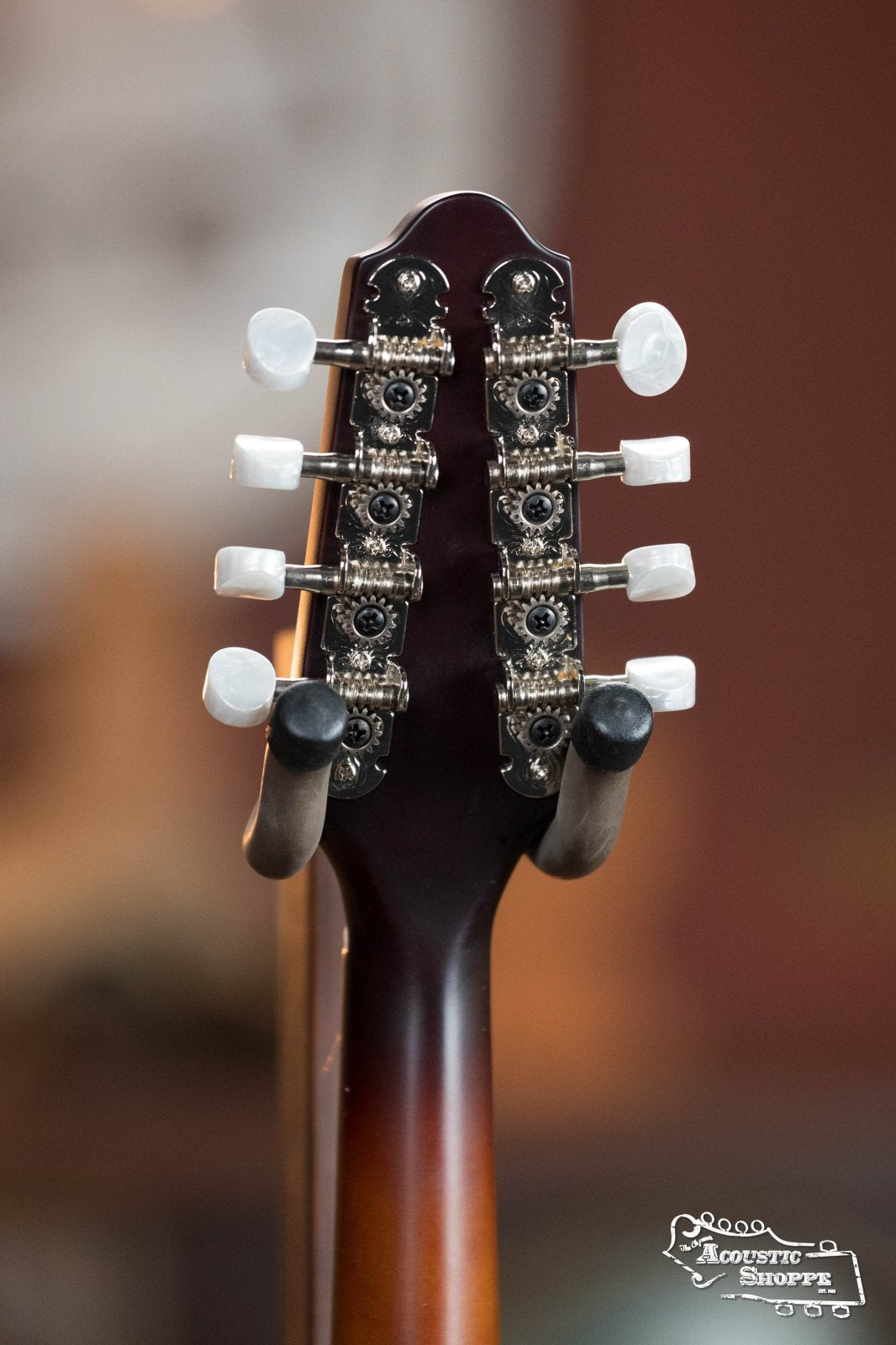 Close-up of the back of The Loar LM-110E-BRB "Honey Creek" A-Style Brownburst Mandolin headstock with tuning pegs, on a stand. The Loar and Acoustic Shoppe logos are visible in the bottom right corner.