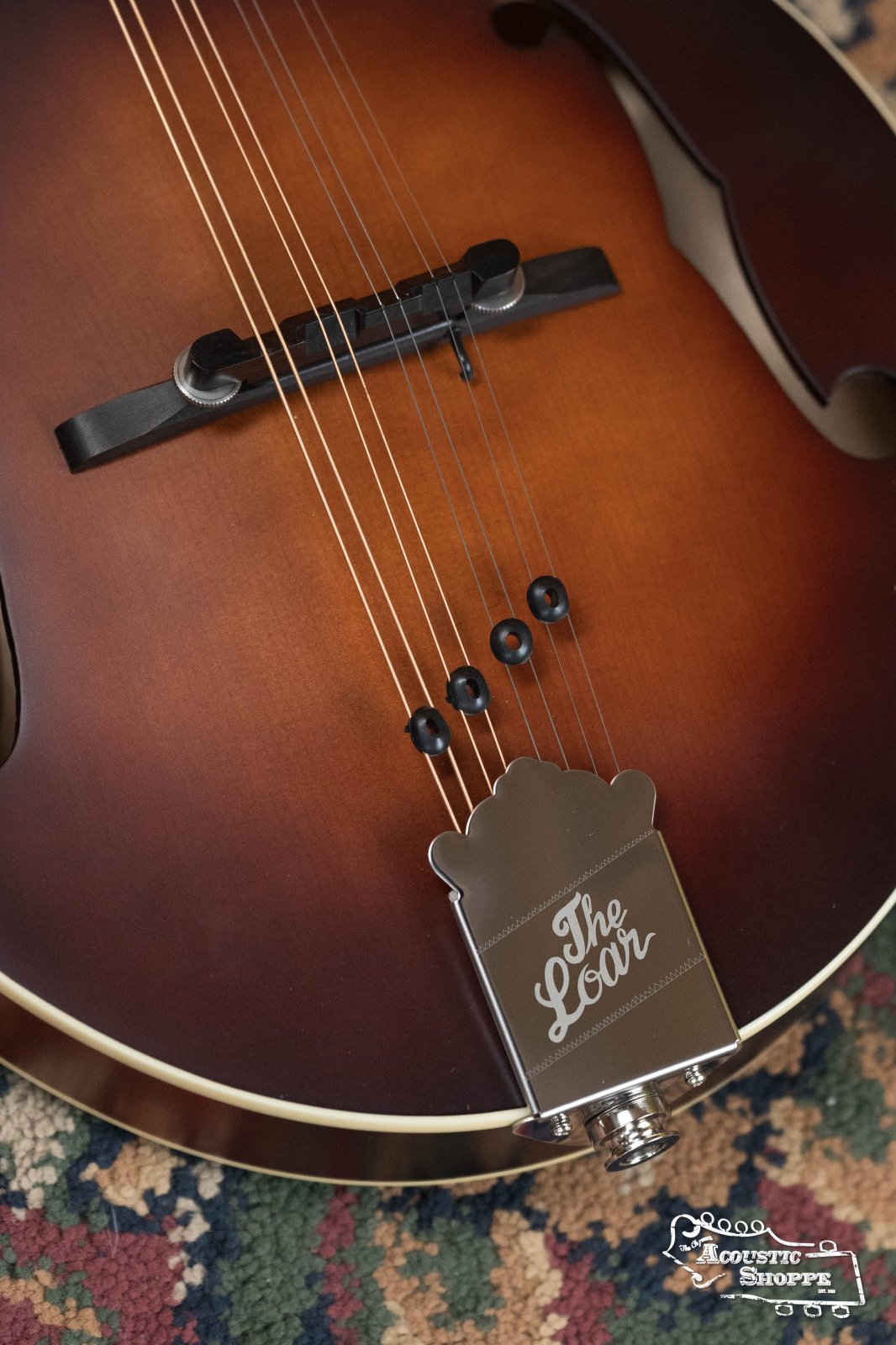 A close-up of The Loar LM-110E-BRB "Honey Creek" A-Style Brownburst Mandolin w/Pickup, showing the bridge, strings, and tailpiece engraved with The Loar, resting on a patterned rug. The Acoustic Shoppe logo appears in the corner.