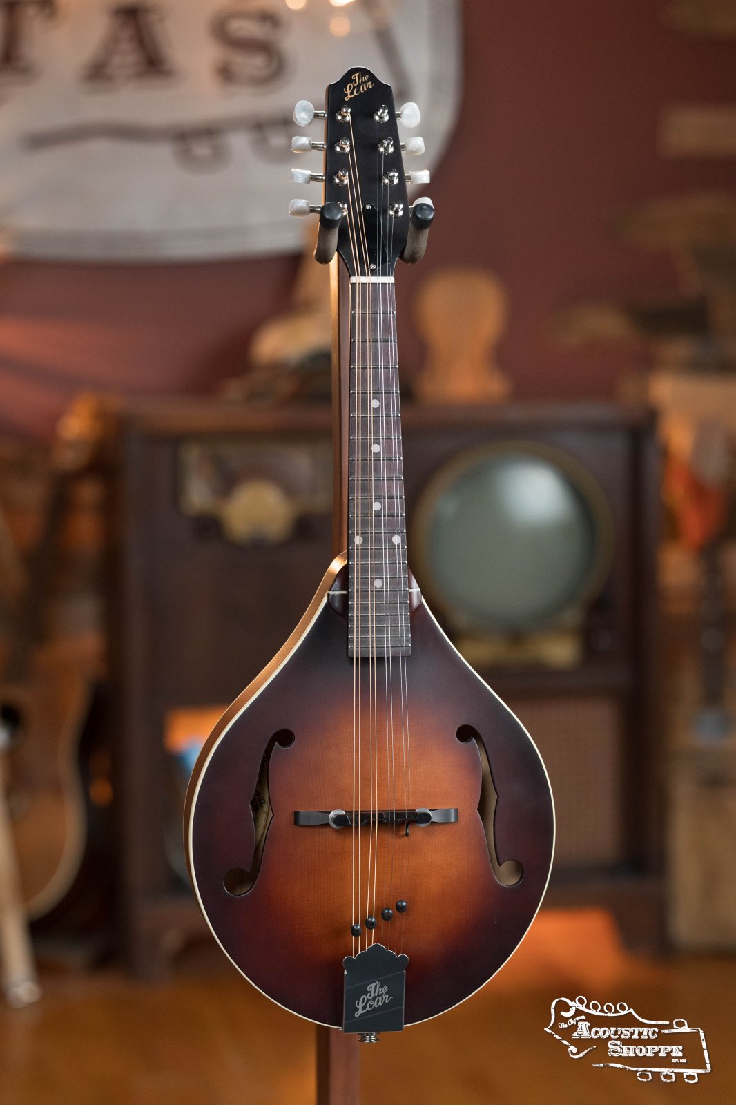 The Loar LM-110E-BRB "Honey Creek" A-Style Brownburst Mandolin with pickup stands upright on a display stand, set against vintage furniture that evokes a warm, nostalgic atmosphere.