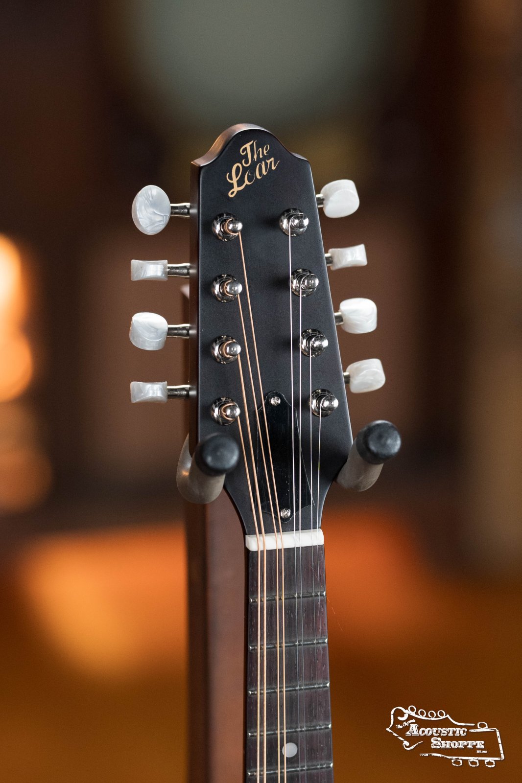 Close-up of the headstock on The Loar LM-110E-BRB "Honey Creek" A-Style Brownburst Mandolin w/Pickup #0688, showing tuning pegs and strings. The Acoustic Shoppe logo appears in a softly blurred background, highlighting its solid spruce top craftsmanship.