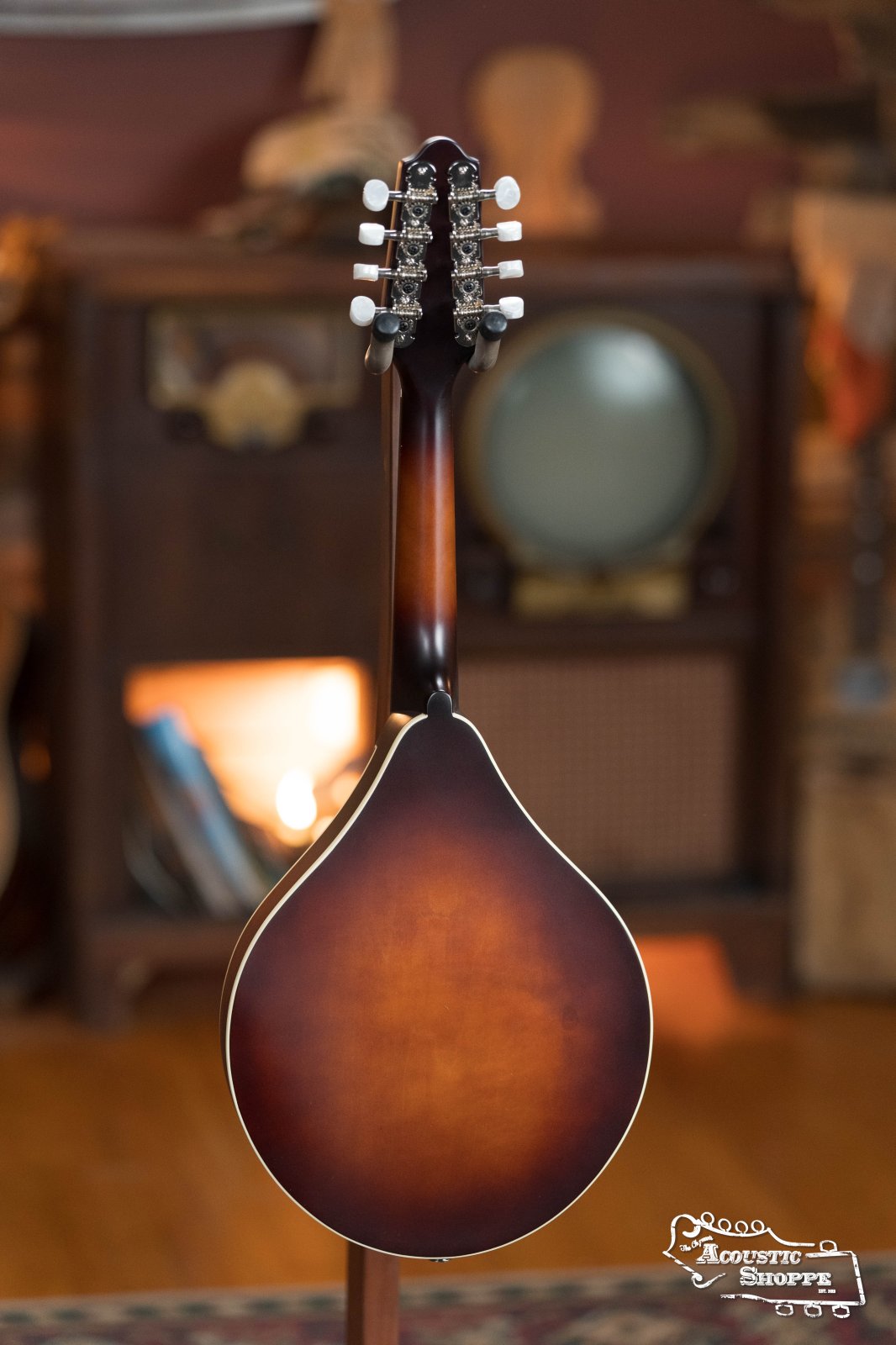 The Loar LM-110E-BRB "Honey Creek" A-Style Brownburst Mandolin w/Pickup #0688 is displayed from the back before a vintage TV and wooden bookshelves, warmly lit with the Acoustic Shoppe logo in the bottom right corner.