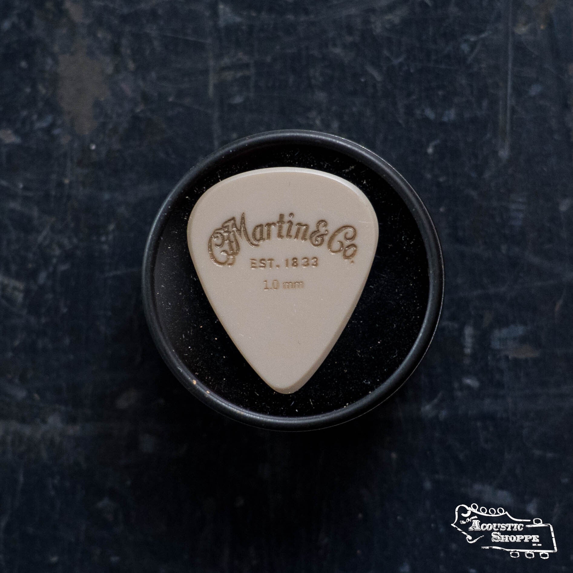 The LUXE By Martin Apex Pick - 1.0mm, a tan, wear-resistant polymer guitar pick from Martin & Co., stands upright in a round black container on a dark textured surface. The Acoustic Shoppe logo appears in the lower right corner.