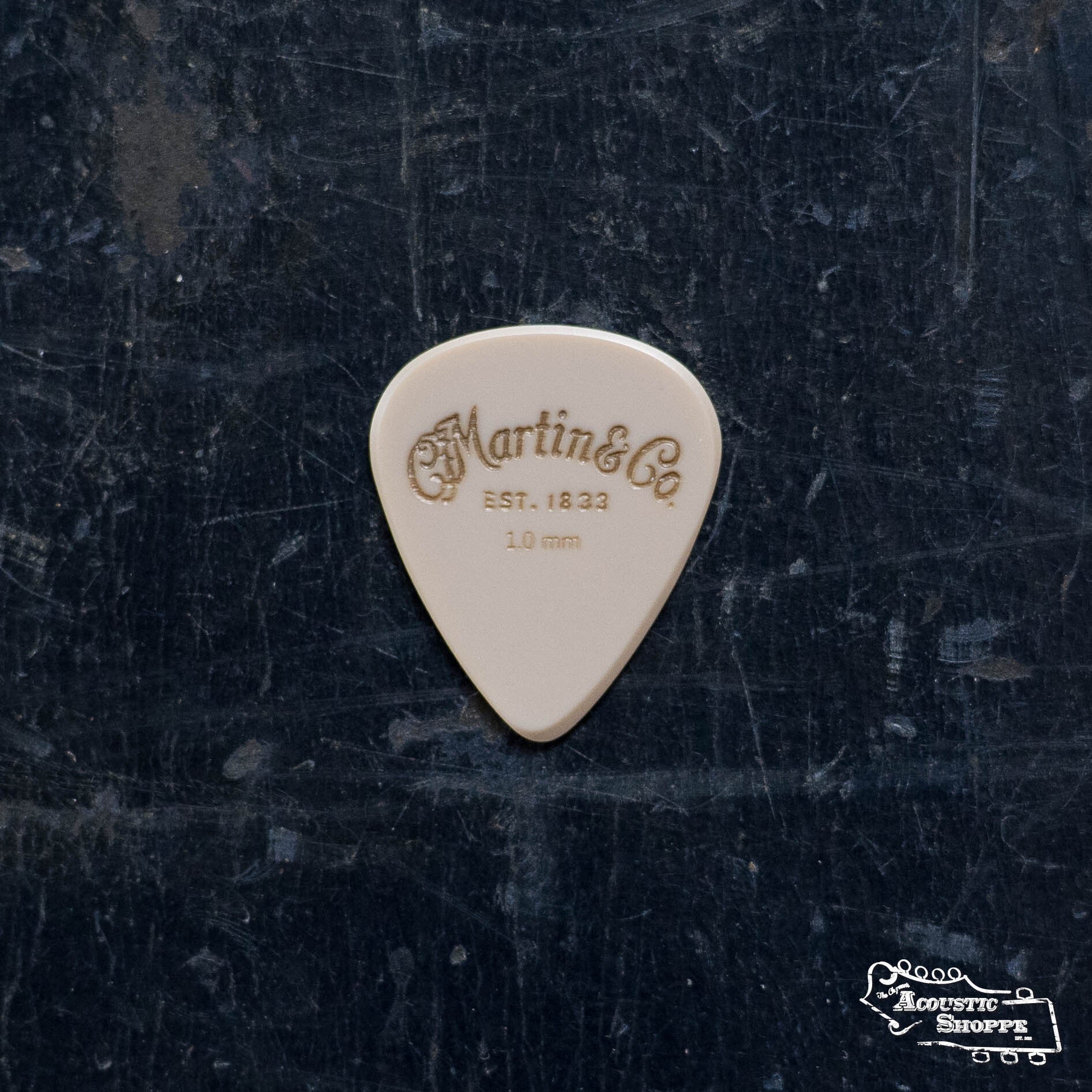 A beige LUXE By Martin Apex Pick - 1.0mm by Martin & Co., made from durable polymer with gold lettering, lies on a dark textured surface. The Acoustic Shoppe logo is visible in the lower right corner.