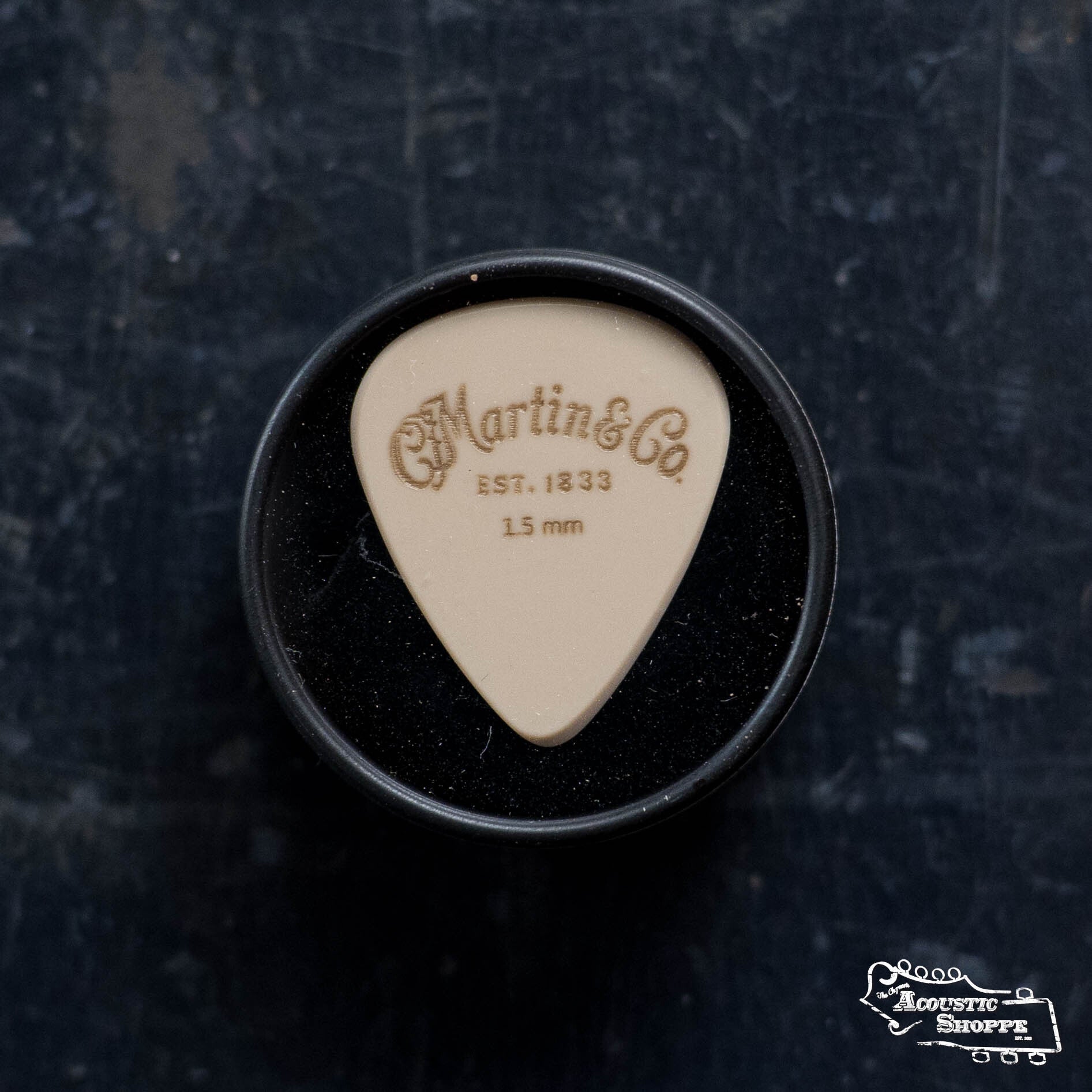 The LUXE By Martin Apex Pick - 1.5mm by Martin & Co., a beige wear-resistant polymer pick with machined bevels, sits in a round black case on a dark surface, The Acoustic Shoppe logo at the lower right.